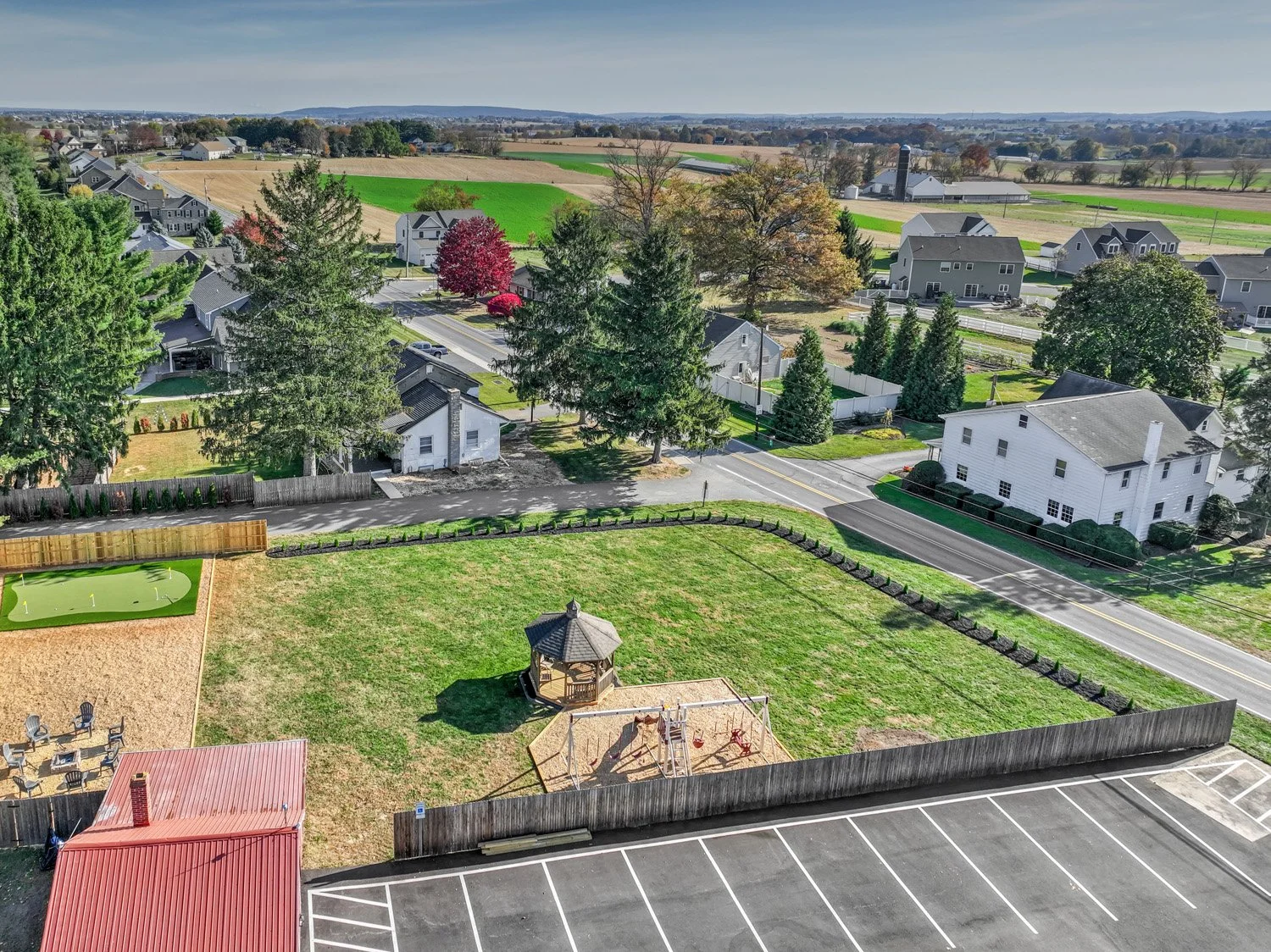 Aerial view of a residential neighborhood with houses, trees, and green fields, along with a fenced yard with a gazebo, sandbox, and playground on a clear day.