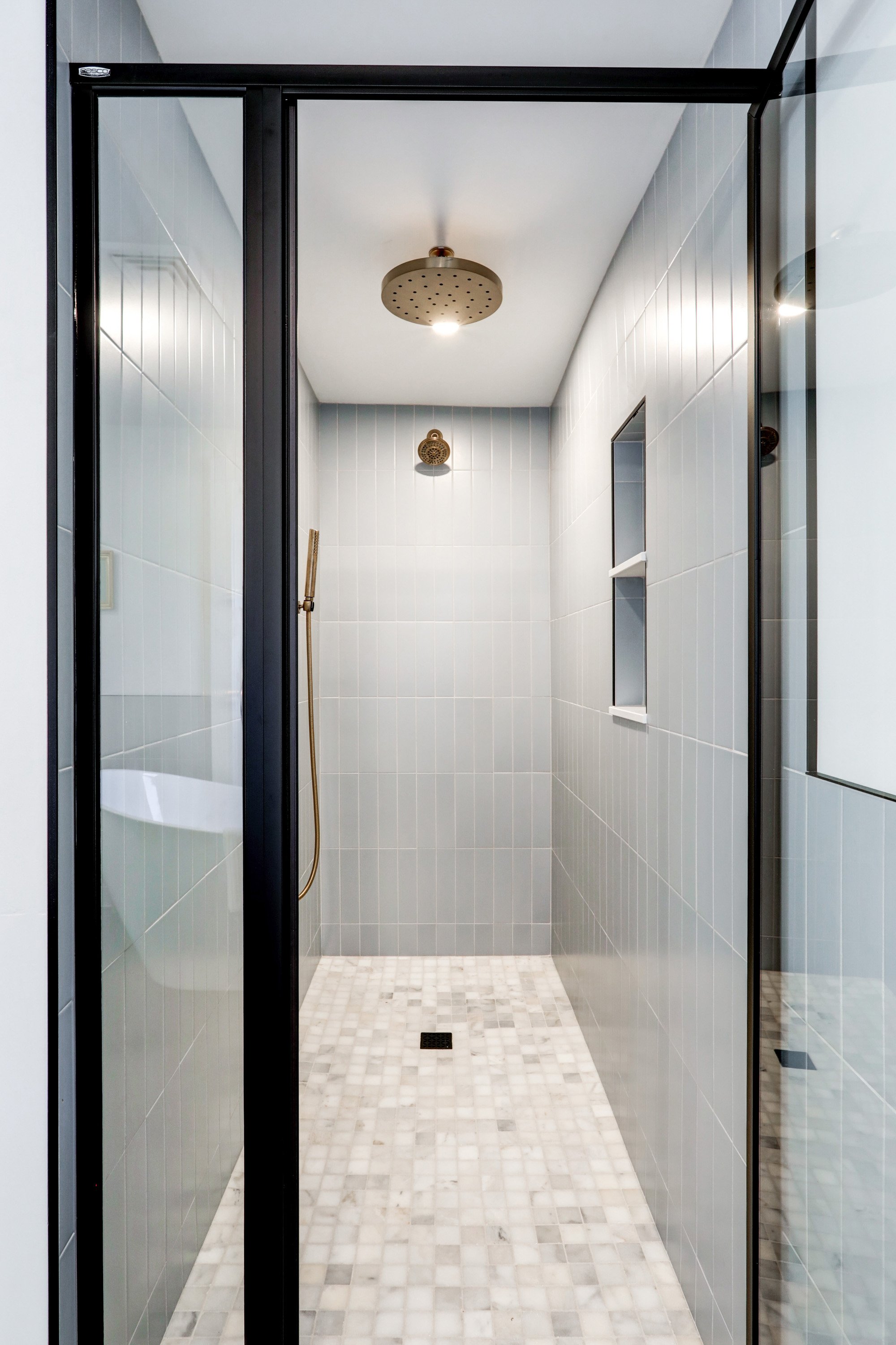 View of a modern shower with glass door, white tile walls, and small square marble floor tiles, featuring a rain shower head and a handheld shower wand.