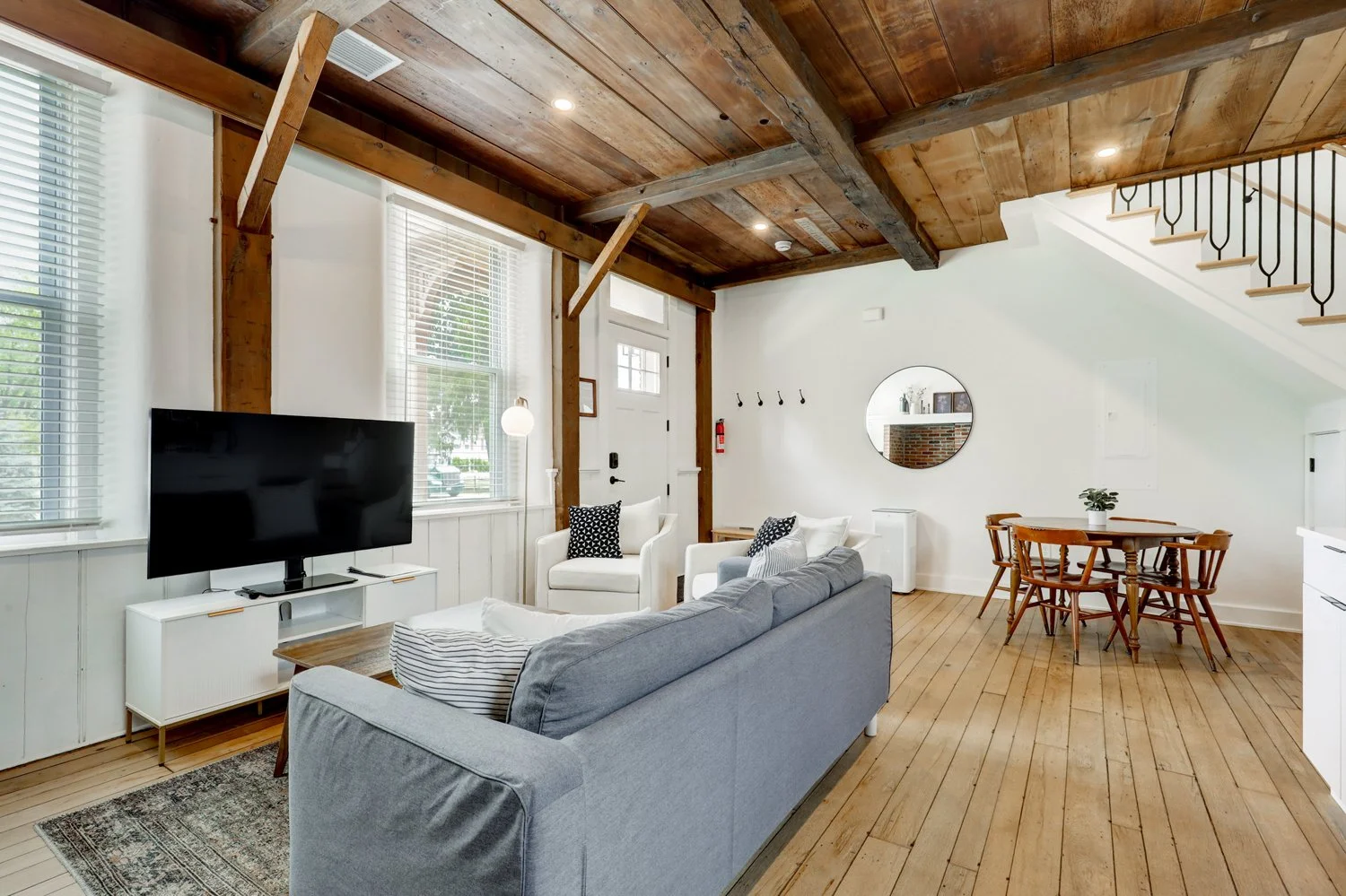Living room with wooden ceiling and beams, white walls, and hardwood floor. Contains a gray sofa, white armchair, black and white pillows, a flat-screen TV on a white stand, a round mirror on the wall, a wooden dining table with four chairs, and a st