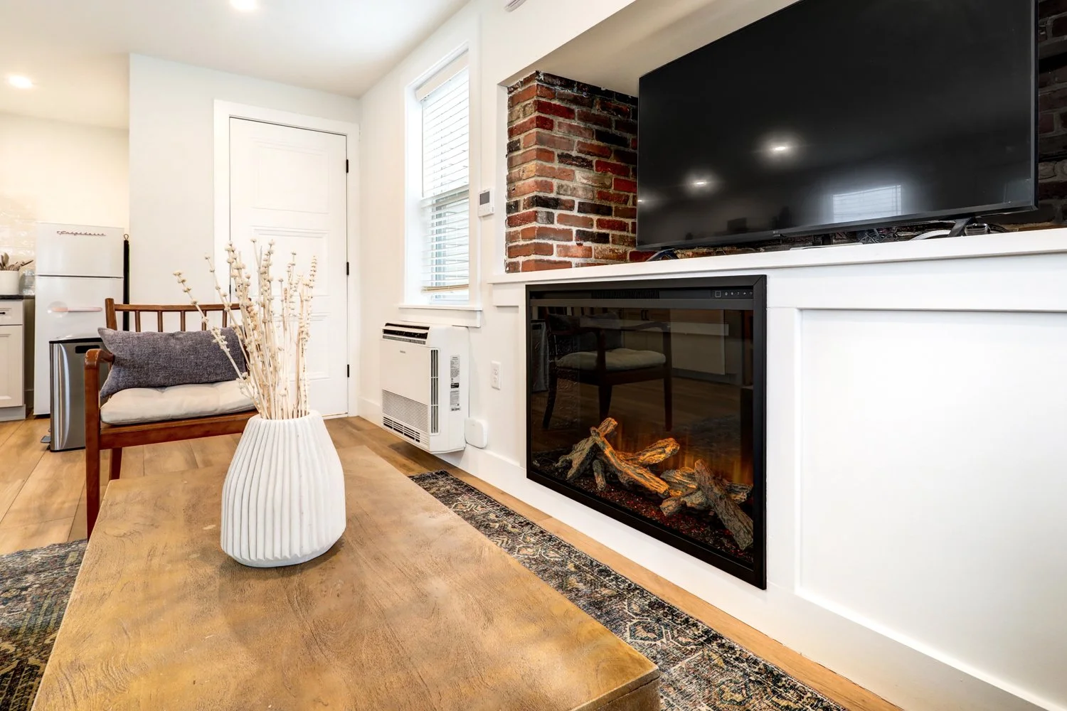 Living room with a white fireplace, black TV, wooden table with ceramic vase, gray cushion on wooden chair, and exposed brick wall