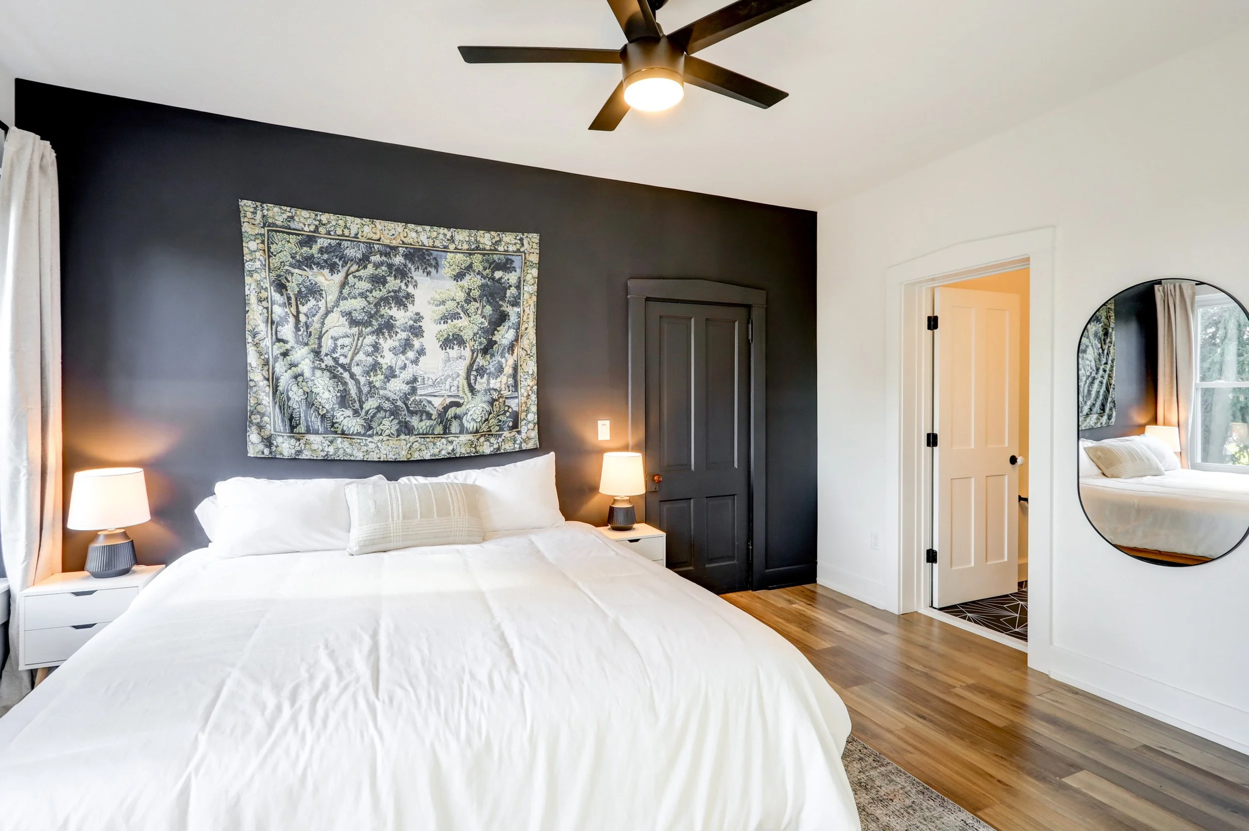 A bedroom with a white bed, black accent wall, and a tapestry with a natural scenery hanging above the bed. There are two nightstands with lamps, a closed black door, a doorway leading to another room, and a round mirror on the wall. A ceiling fan wi