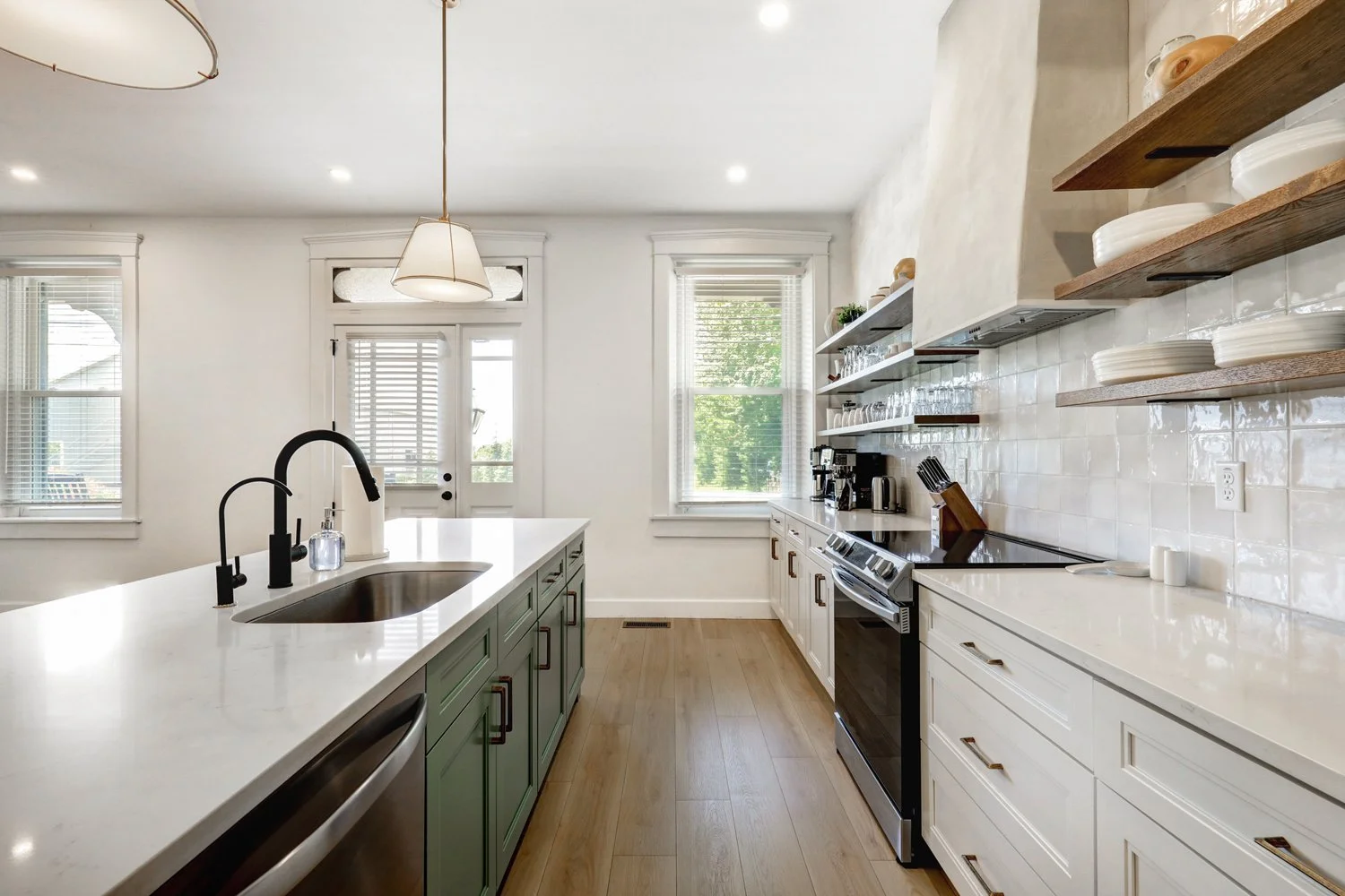 Bright kitchen with white walls, wooden flooring, open shelves with dishes, a white countertop, green cabinets, and a black faucet.