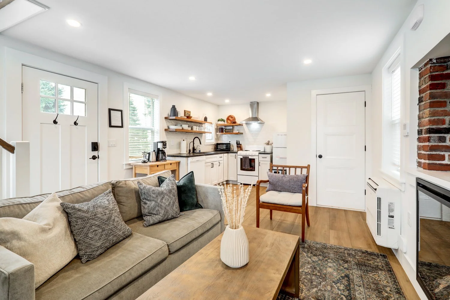 Open-concept living room and kitchen with beige sofa, wooden coffee table, decorative pillows, open shelves, white cabinets, and small kitchen appliances.