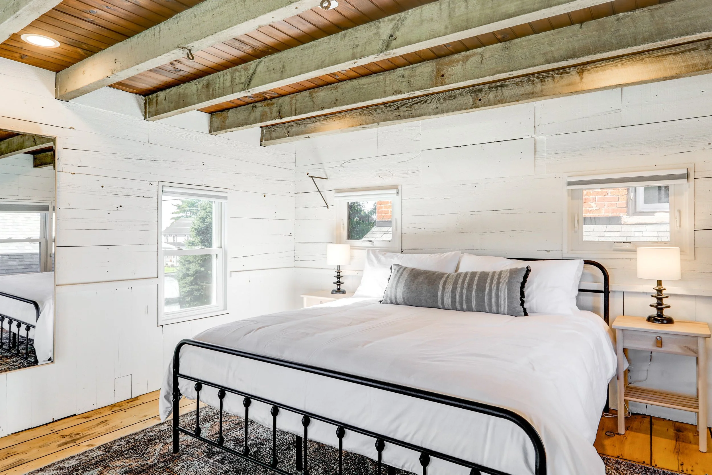 A bedroom with white wooden walls and ceiling beams, a bed with white bedding and gray pillows, two small windows, and a wooden nightstand with a lamp.