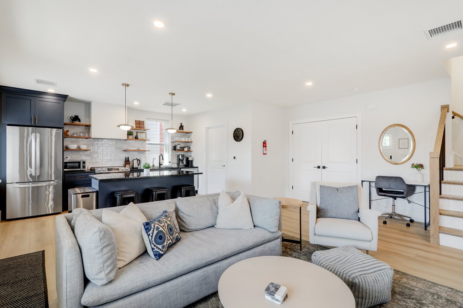 Living room with beige sofa, armchair, and ottoman, open kitchen with stainless steel appliances, black cabinets, open shelves, and island with barstools, hardwood floors, and white walls.