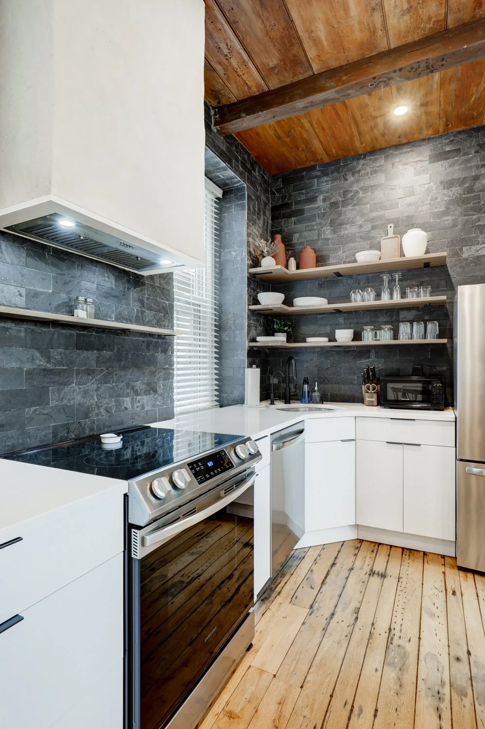 Modern kitchen with dark gray stone backsplash, white cabinets, wooden open shelves with dishes and glassware, stainless steel appliances, and wooden ceiling and floor.