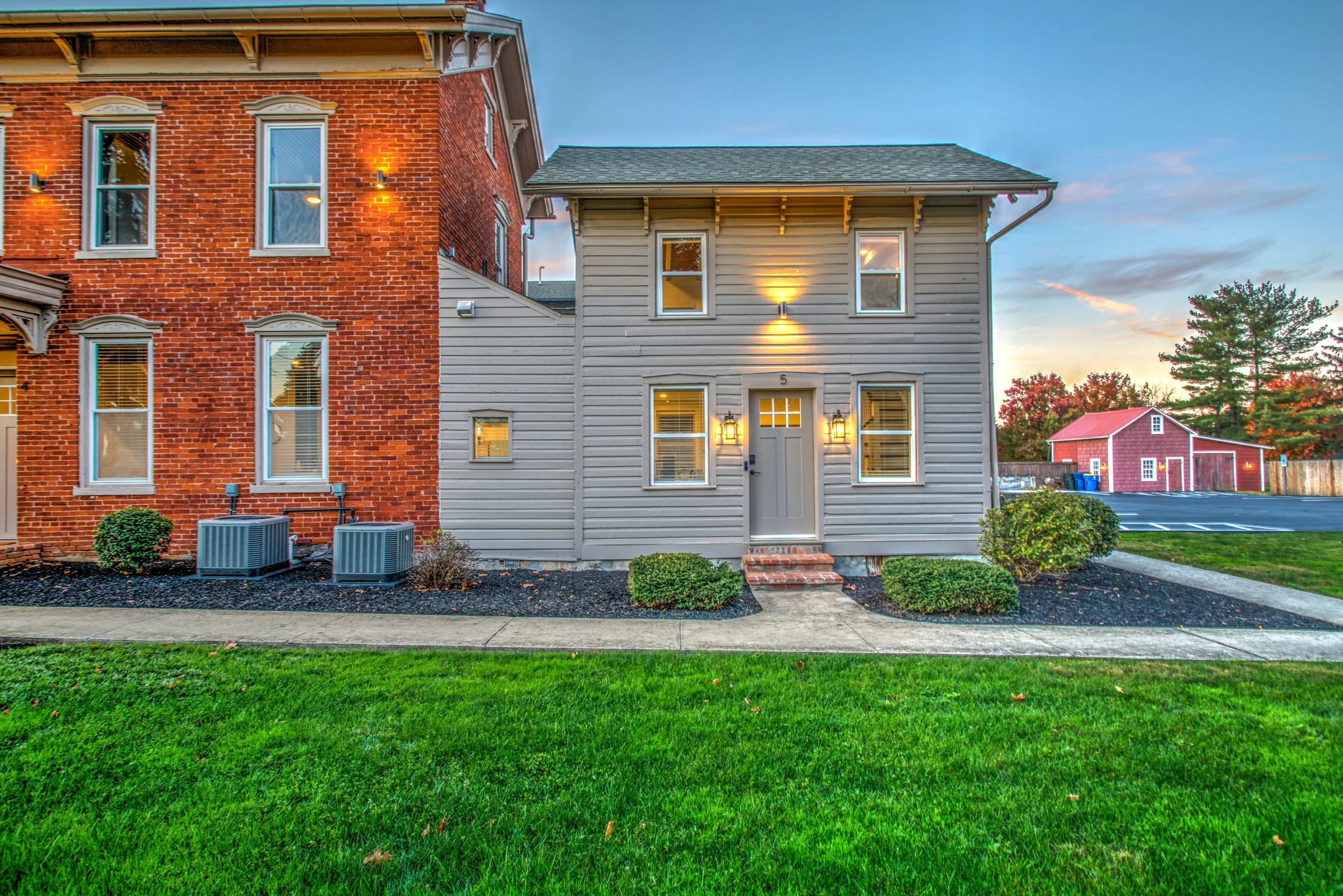 A two-story house with a gray wooden exterior and a brick building attached on the left. The house has multiple windows with white trim, outdoor lighting, and a front door with lantern-style lights. There is a manicured lawn with small bushes, a side