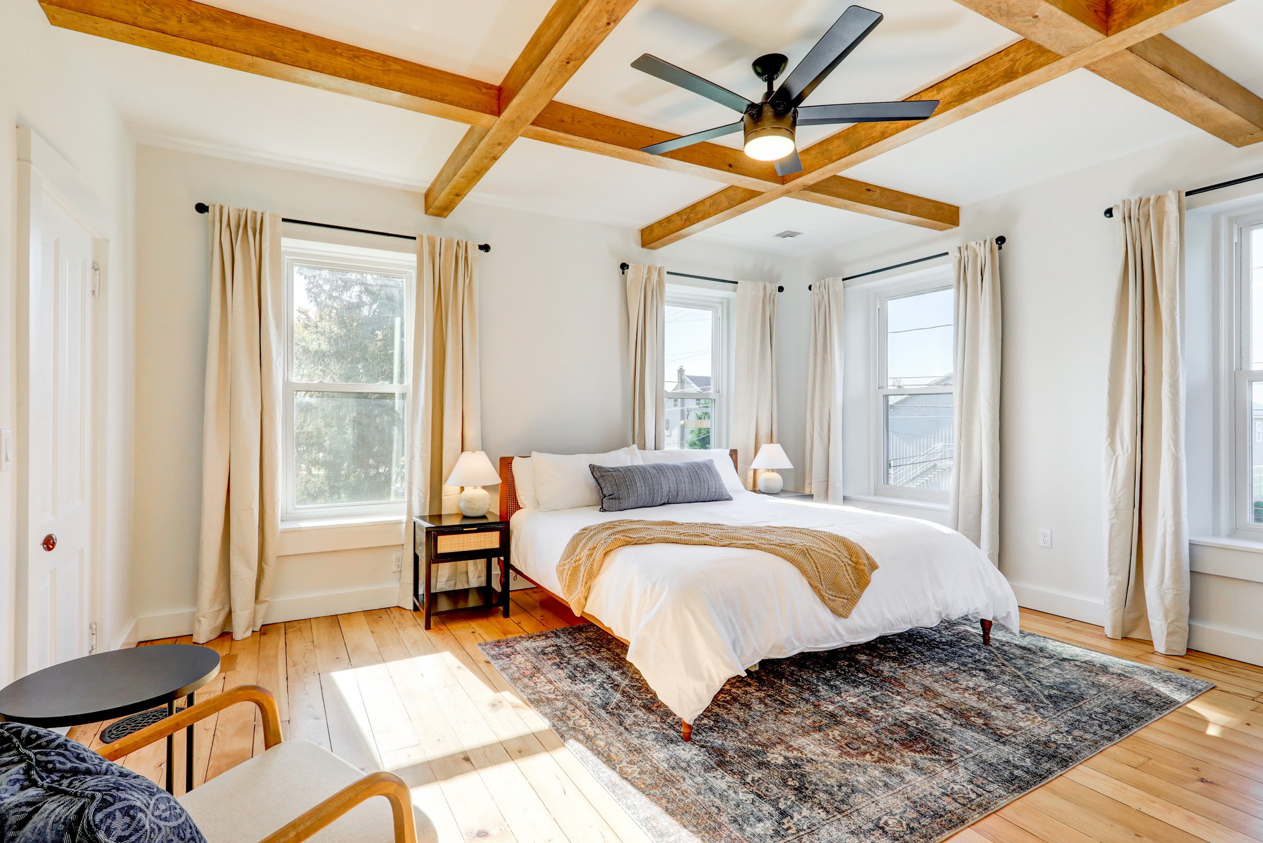 Bright bedroom with a wooden beam ceiling, a large bed with white linens and a gray pillow, two nightstands with lamps, and multiple windows with cream curtains, sunlight streaming in.