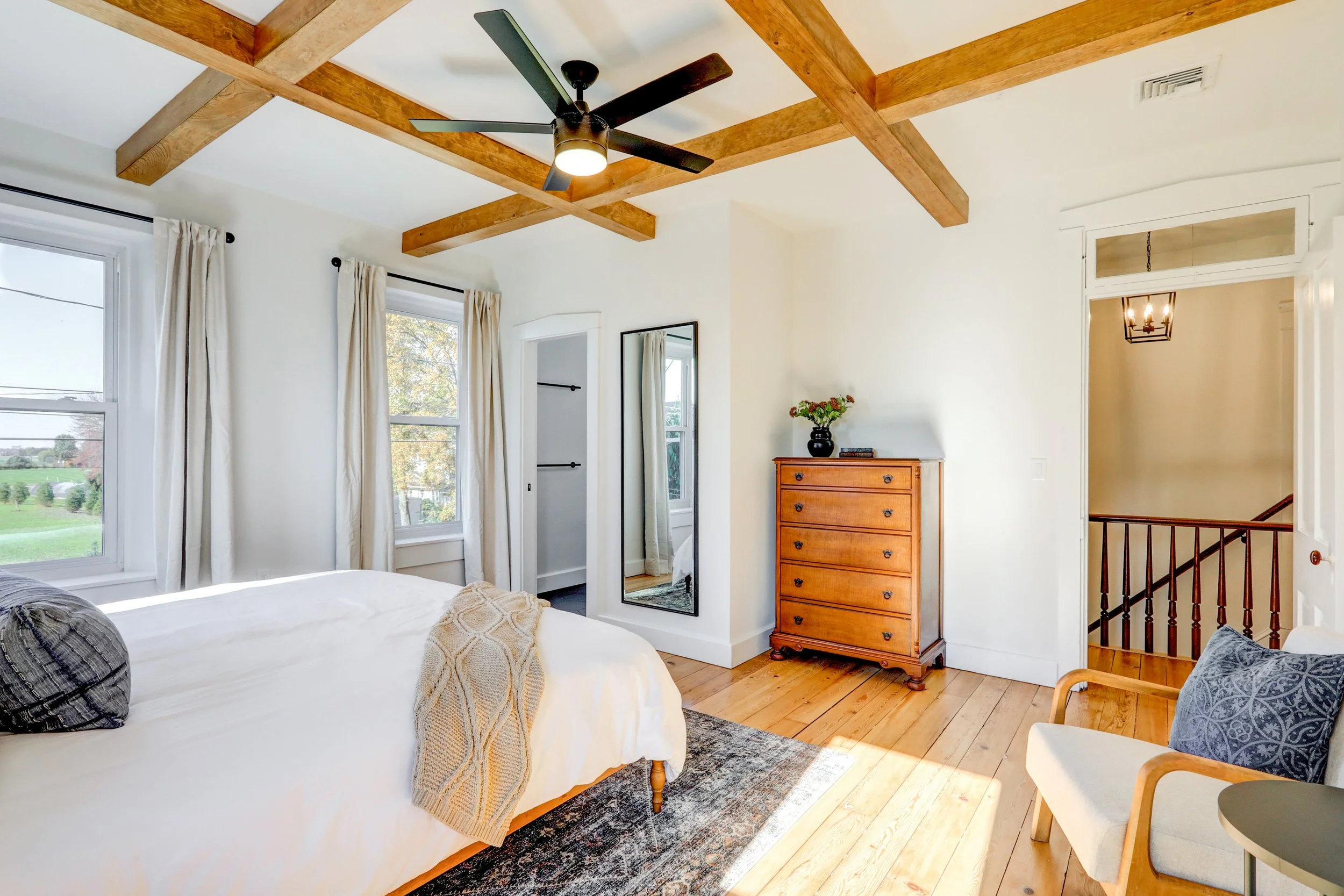 A bedroom with white walls, two windows with white curtains, a wooden dresser with a black vase of flowers, a large mirror, a bed with gray pillows and a beige blanket, hardwood floors, and a ceiling with wooden beams and a modern ceiling fan.