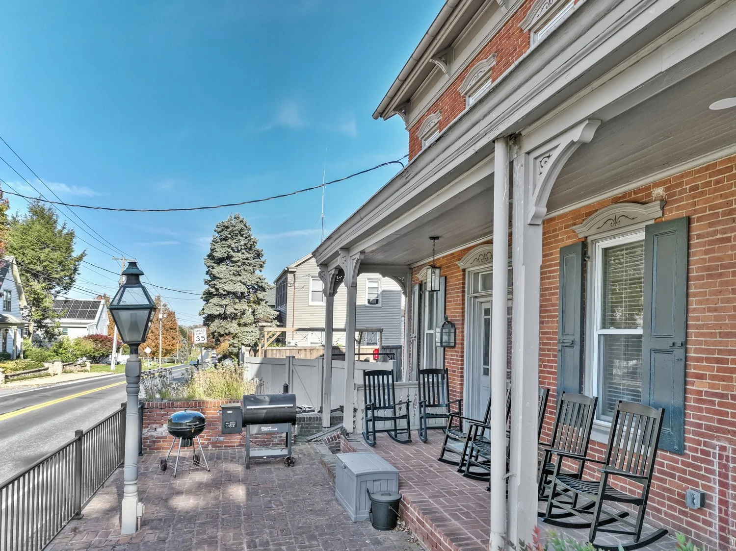 Porch of a brick house with several black rocking chairs, a mailbox, a barbecue grill, and a small table. Street with houses, trees, and a blue sky in the background.