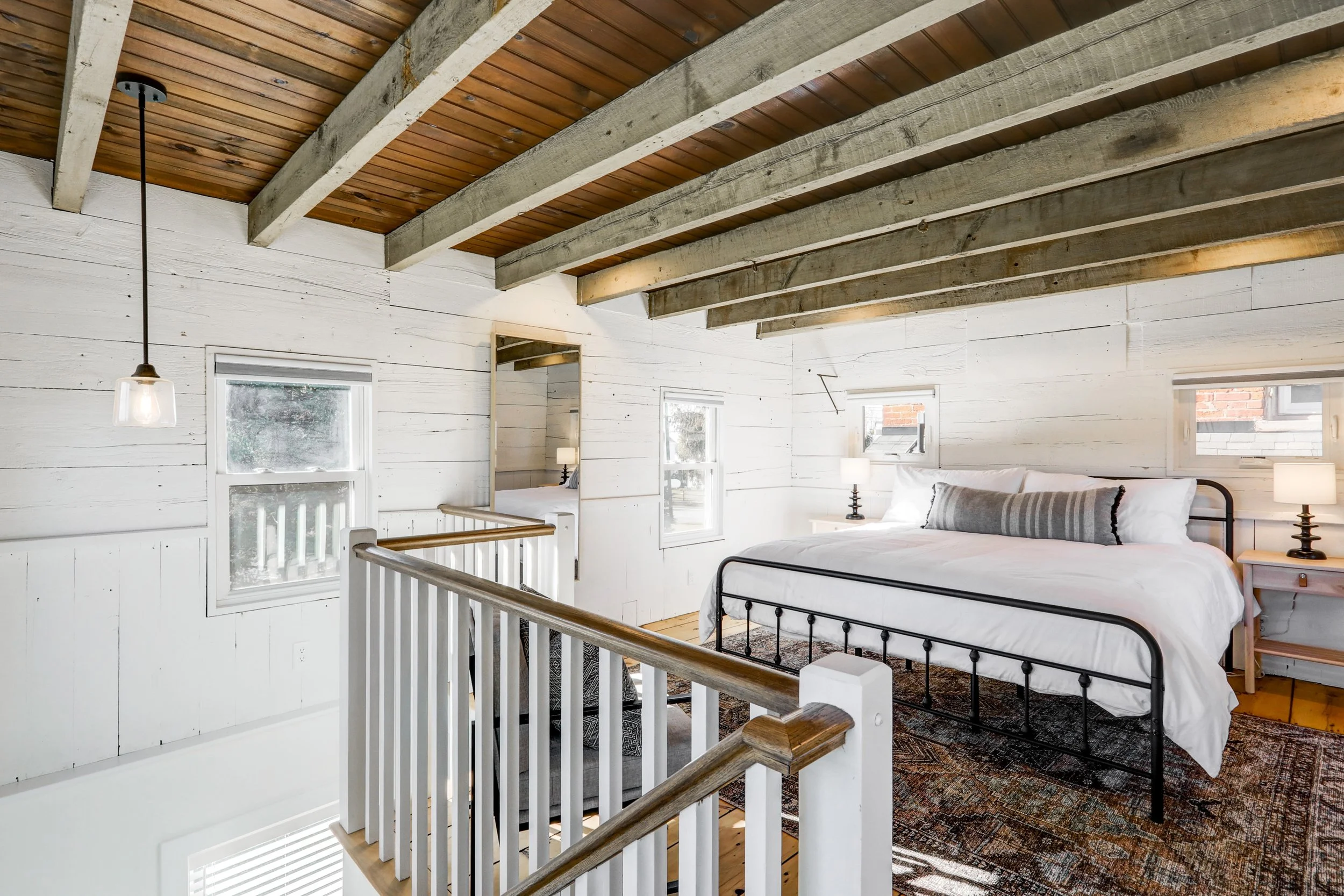 Loft bedroom with white wooden walls, exposed ceiling beams, and a bed with white bedding and a striped pillow, illuminated by natural light from windows.