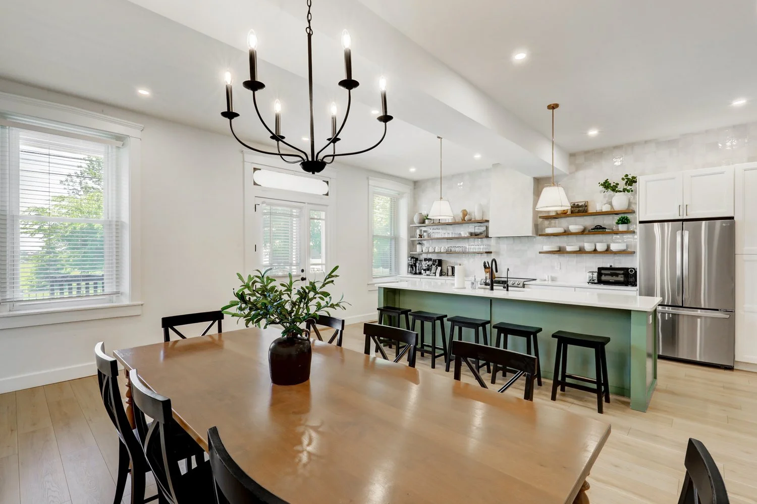 Bright kitchen and dining area with a wooden dining table, black chairs, and a green kitchen island with black stools. White cabinets, open shelves with dishes, a refrigerator, and large windows with blinds.