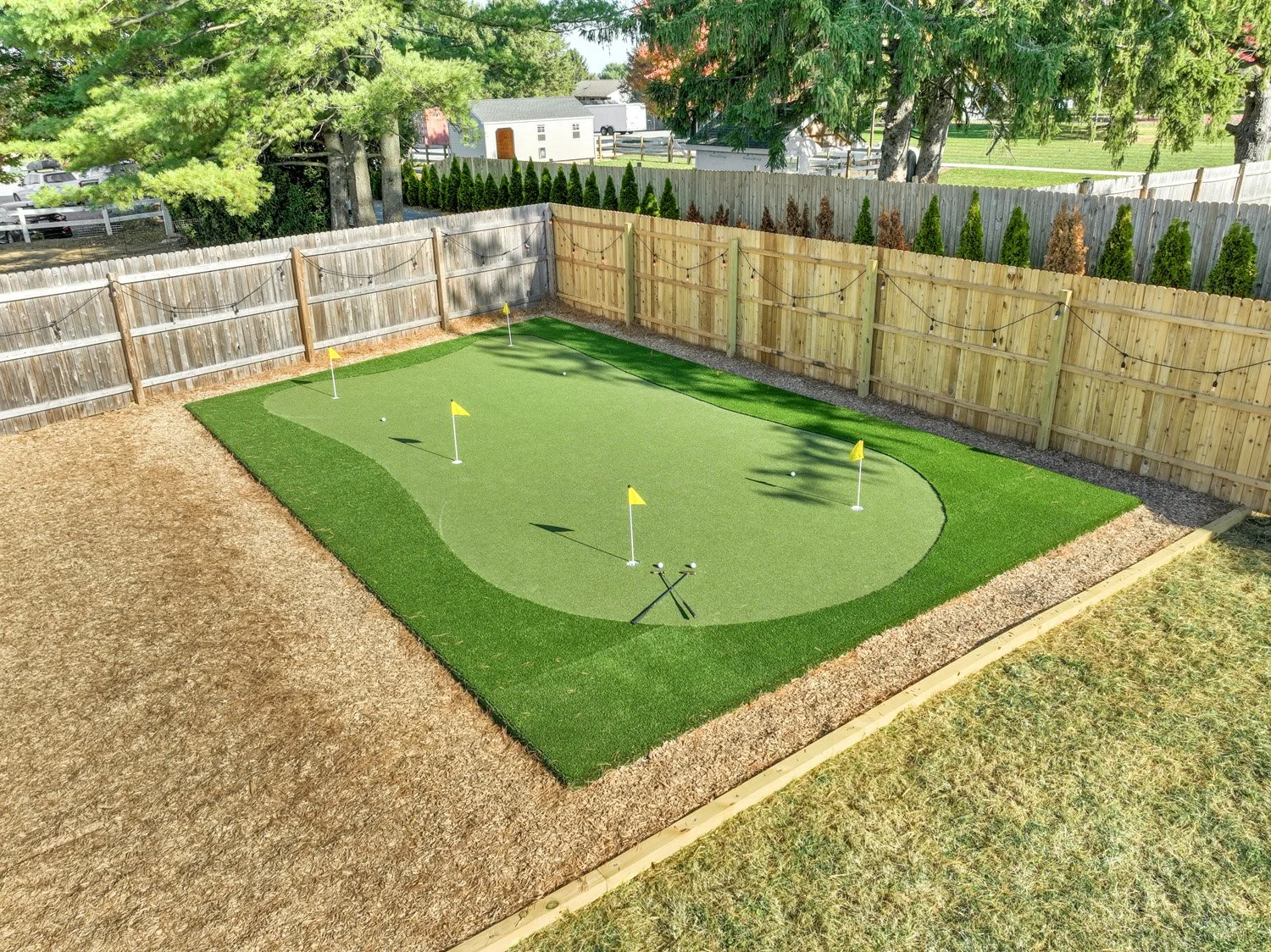Backyard putting green with five flags, golf balls, and golf tees, enclosed by wooden fencing, with trees and neighboring houses visible.