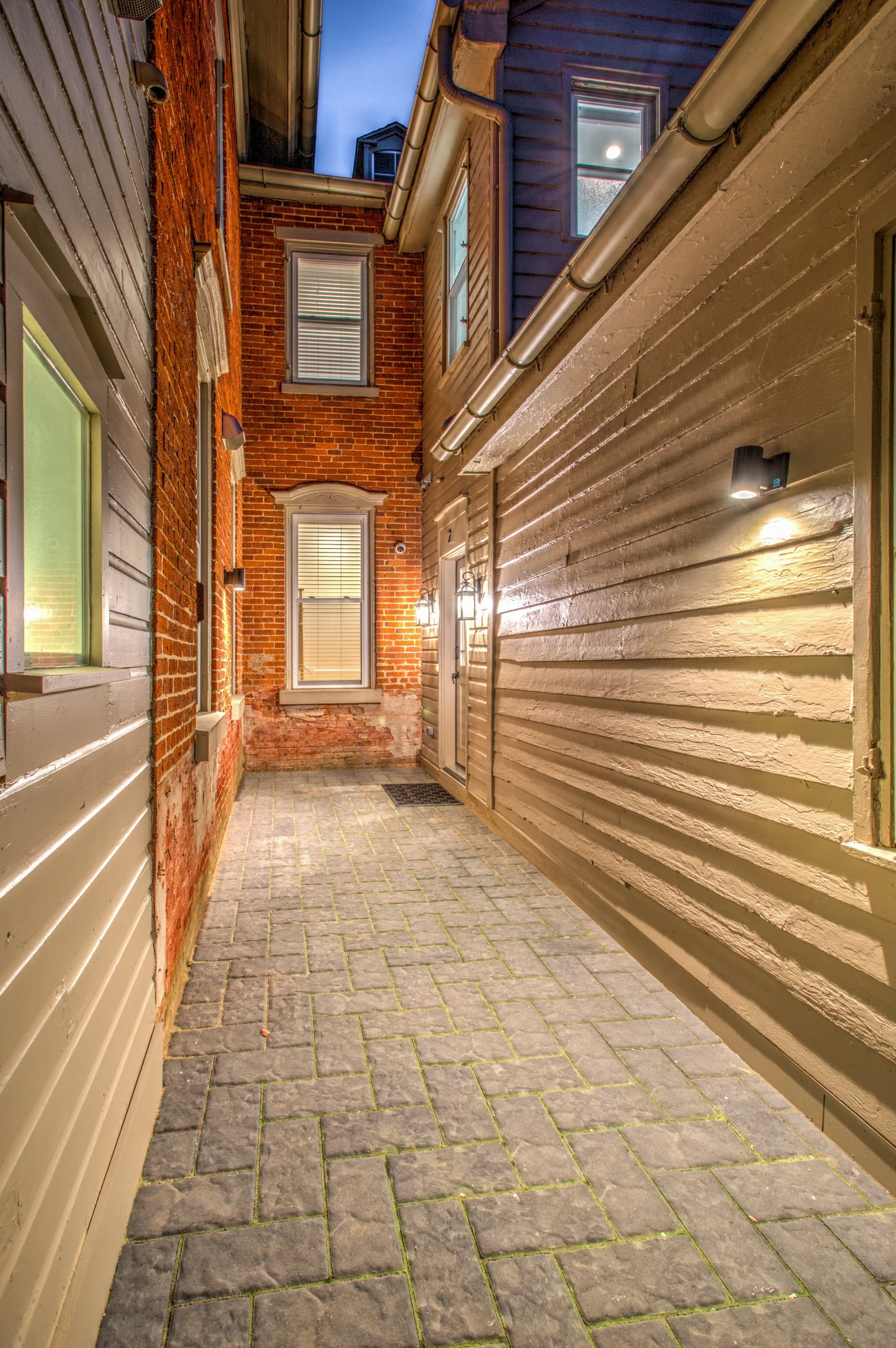 A narrow brick and wooden alleyway with a paved stone floor, illuminated by wall-mounted outdoor lights. The alley is surrounded by buildings with brick, wood, and siding exteriors, with several windows visible. The sky is visible at the top of the i