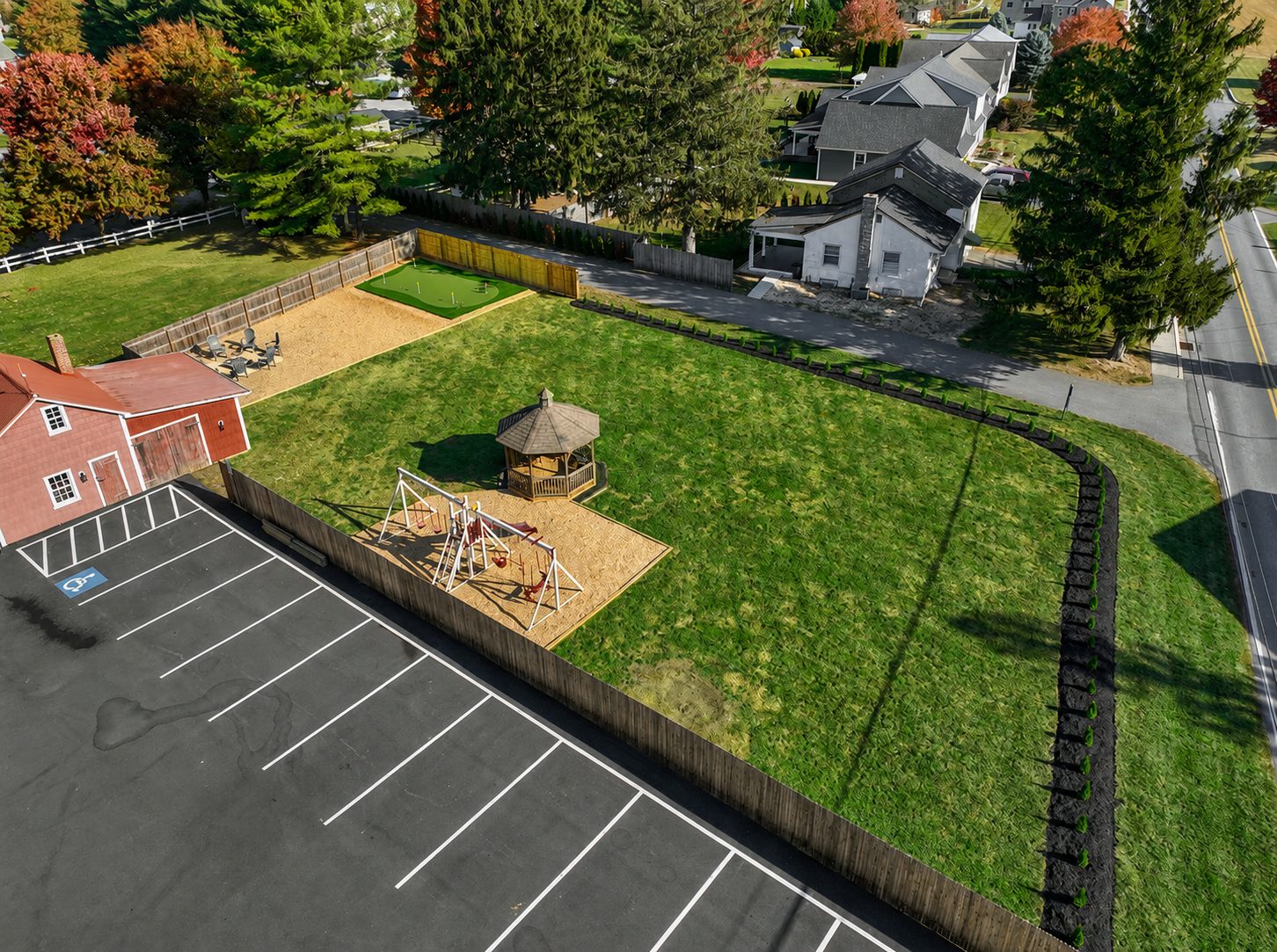 An aerial view of a residential backyard that includes a colorful playground with swings and a small covered gazebo, a sandpit, and a fenced-in putting green, with a parking lot and neighboring houses and trees surrounding the yard.
