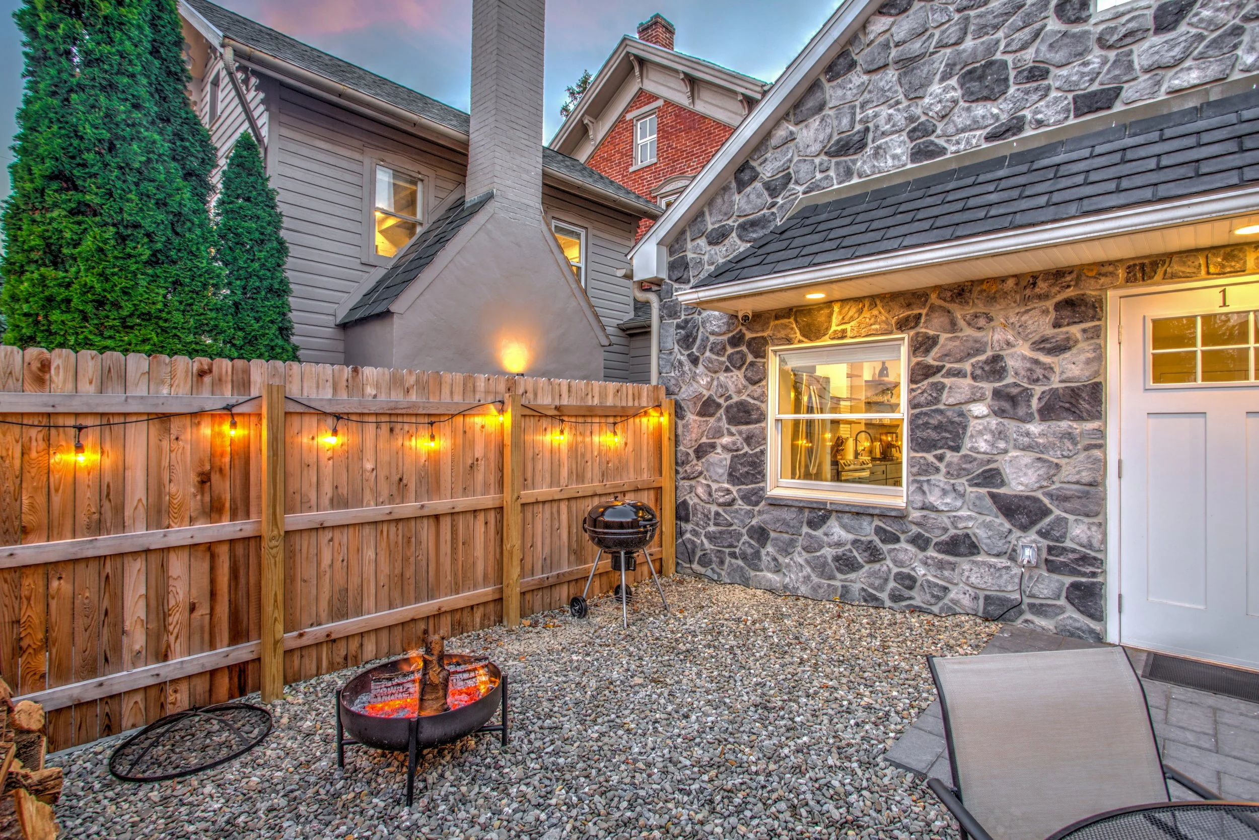 A cozy backyard patio with a wood fence, string lights, a fire pit with glowing embers, a gravel ground, a black grill, and outdoor chairs. The house has stone and siding exterior walls, with lit windows at sunset.