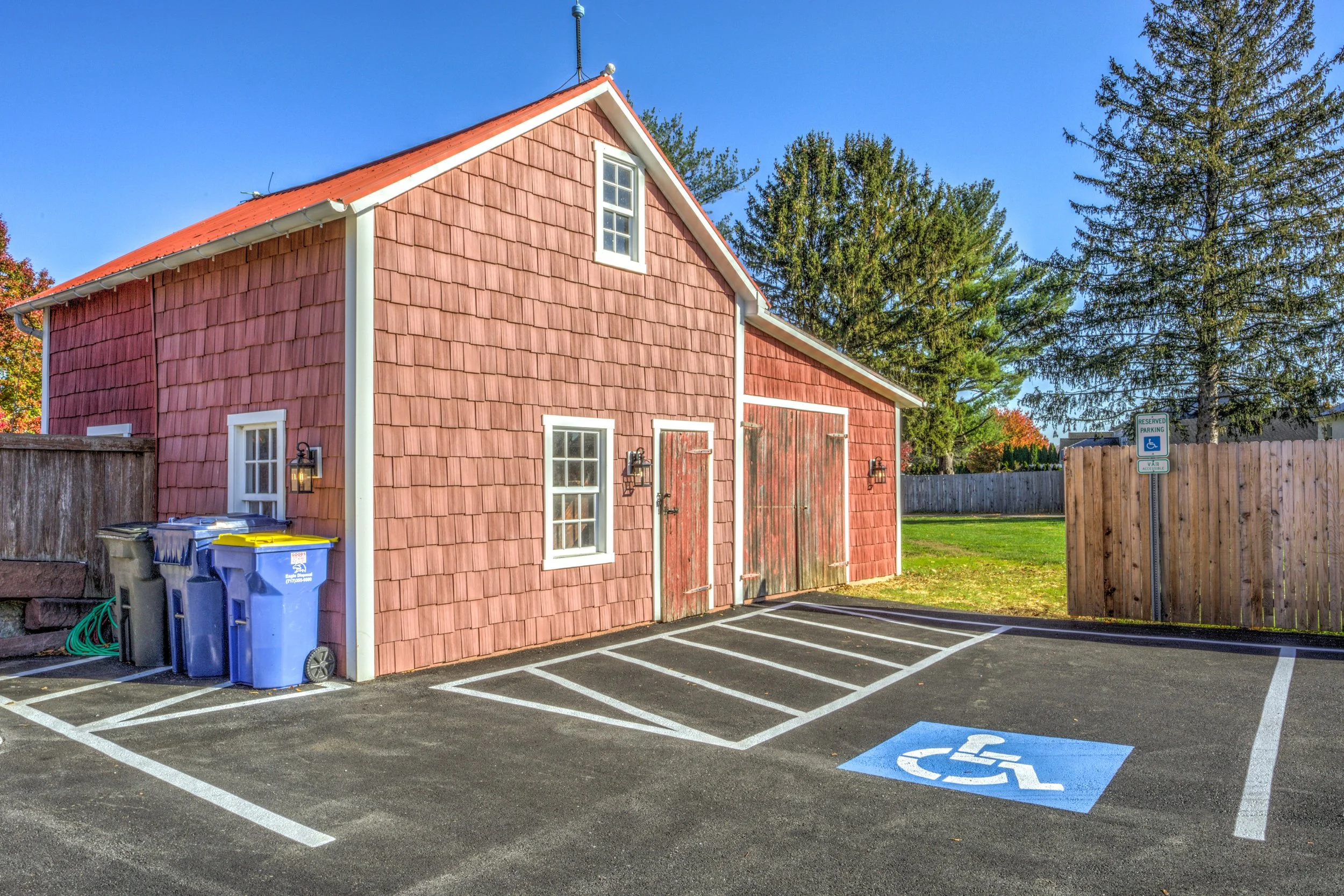 A building with pink wooden siding, white window frames, and a red metal roof, situated in a parking lot with accessible parking space marked for handicapped. There are green trees and a wooden fence in the background.