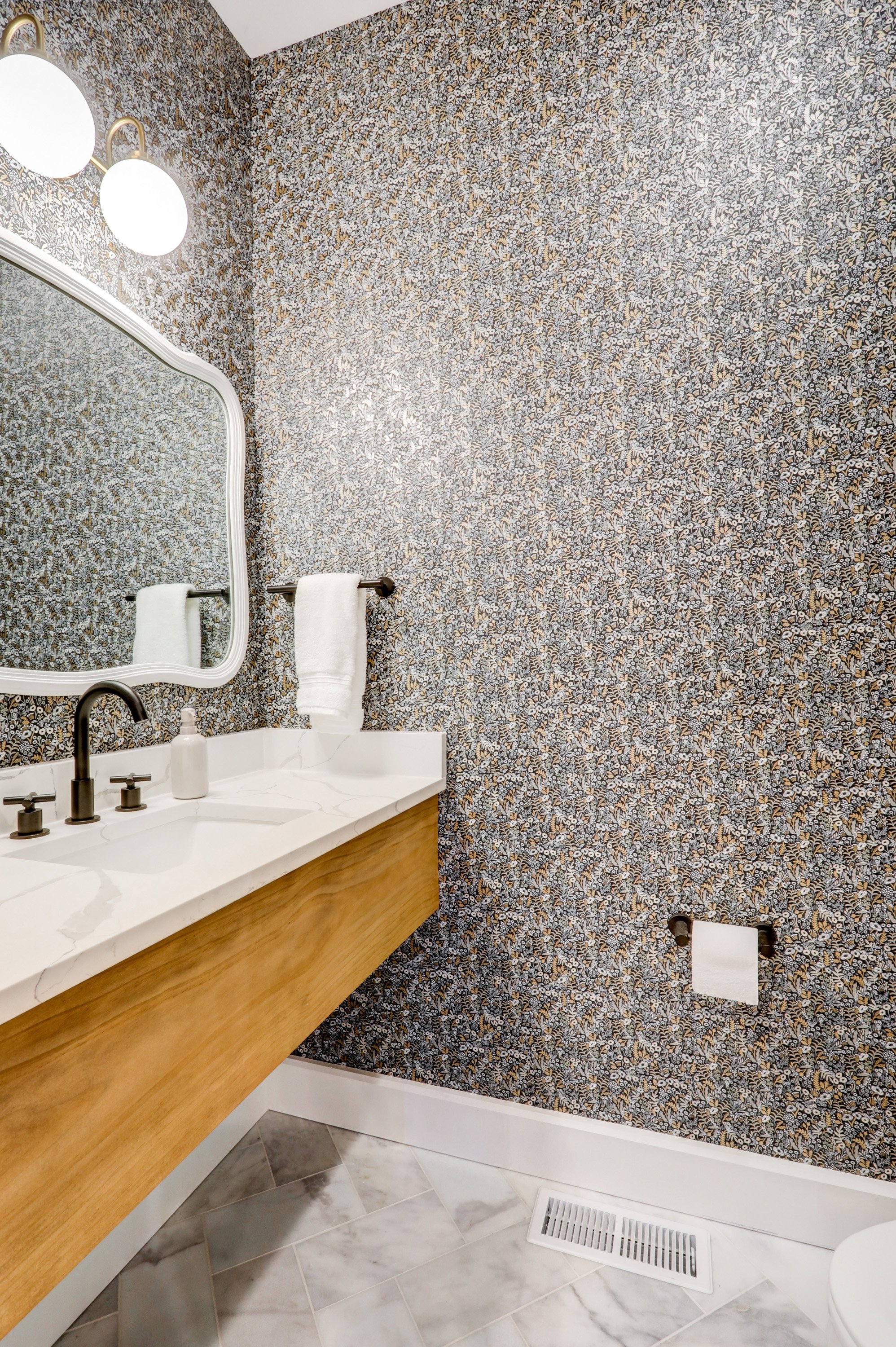 Bathroom with floral wallpaper, white marble countertop with black faucet, mirror, white towel, and toilet paper holder with a white towel, tiled floor with gray and white marble pattern, and floor vent.