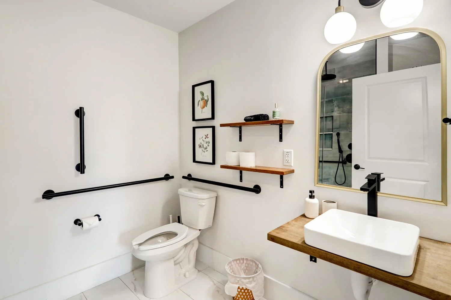 Modern bathroom with white walls, a toilet, black safety rails, wooden shelves, framed artwork, a large mirror, a white sink on a wooden countertop, and a glass shower enclosure in the reflection.