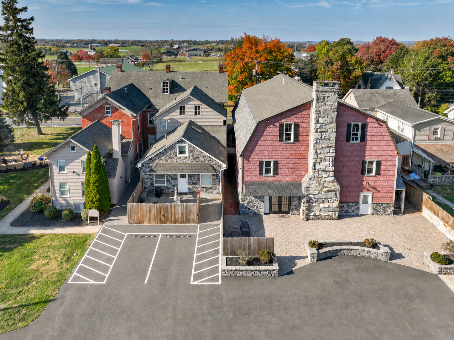 Aerial view of a residential neighborhood with a parking lot in the foreground, surrounded by houses with varied architectural styles and colors, including a prominent pink house with a stone chimney. Trees with fall foliage are visible in the background.