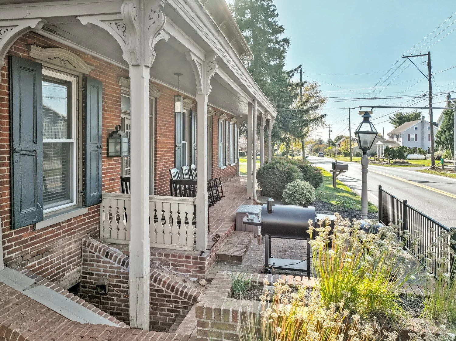 Front porch of a brick house with black shutters, white decorative woodwork, four chairs, a barbecue grill, and a street view with trees and old-fashioned street lamps.