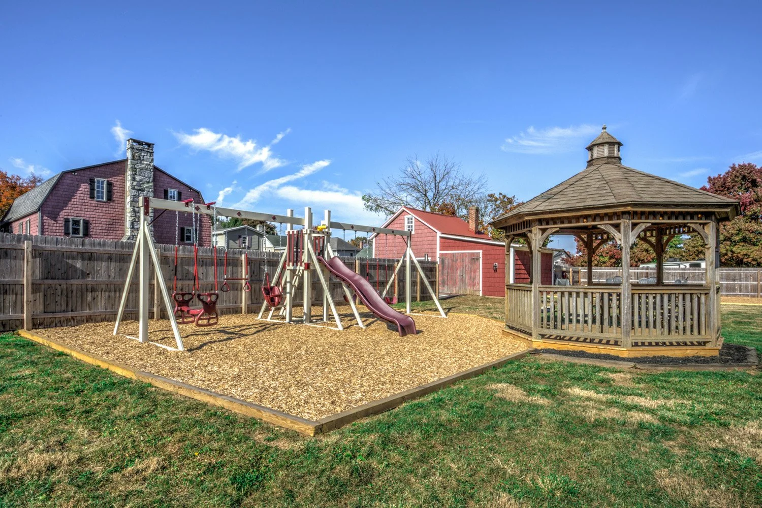 A backyard play area with a wooden sandbox and a swing set that has two swings and a slide, enclosed by a wooden fence, and a wooden gazebo on the right, with a house and trees visible in the background under a blue sky with scattered clouds.