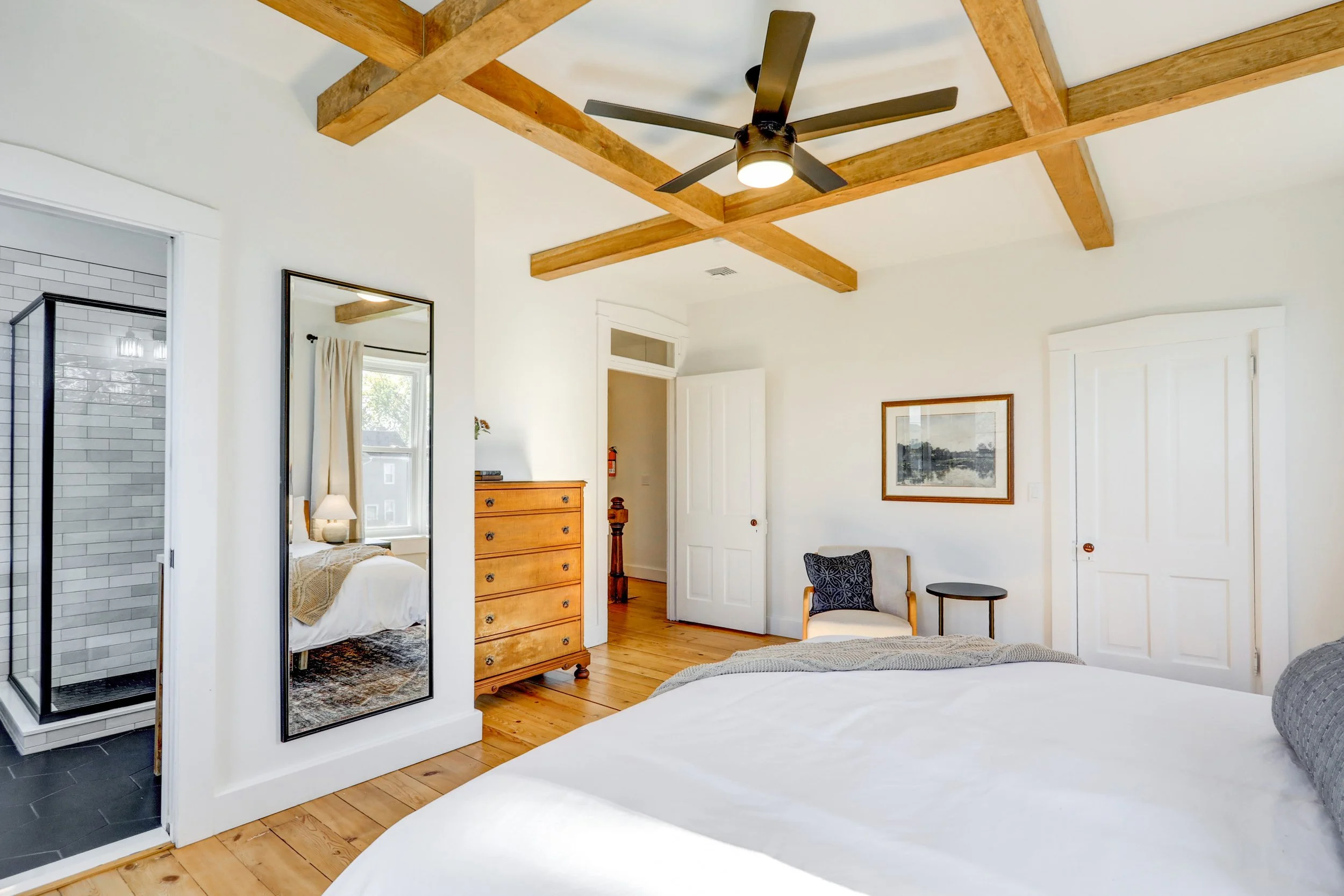 A bedroom with a white ceiling and wooden beams, a ceiling fan, a mirror, a wooden dresser, a window with curtains, a bed with white bedding, a chair, and a small table.
