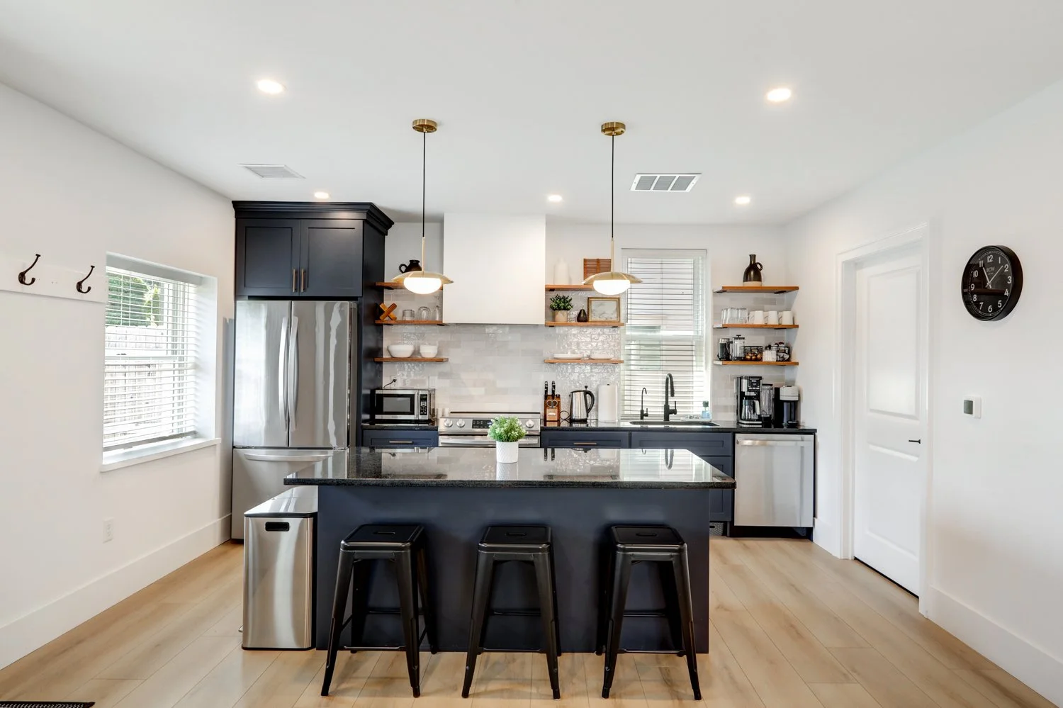 Modern kitchen with navy blue cabinets, stainless steel appliances, a black island with three black stools, open wooden shelves, white and gray backsplash, and hardwood floors.