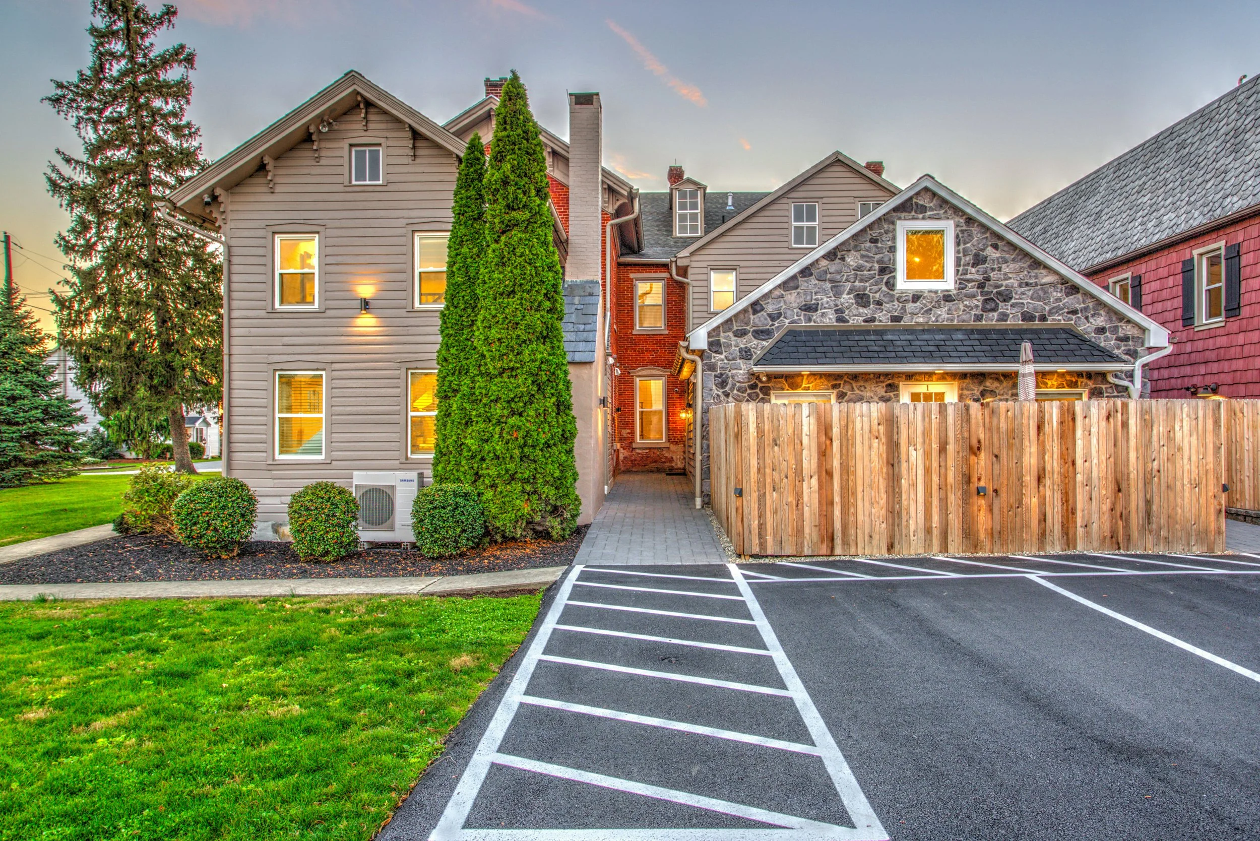 A residential building with multiple styles including siding, stone, and brick, surrounded by greenery and a parking lot with white striped spaces.