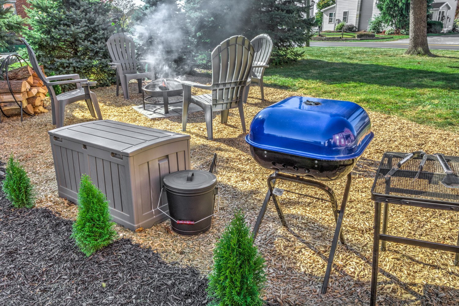 Backyard patio with outdoor furniture, a fire pit with smoke, a blue grill, a storage bench, a trash bin, tools on a table, small bushes, and a woodpile with chopped logs, with grass and trees nearby.