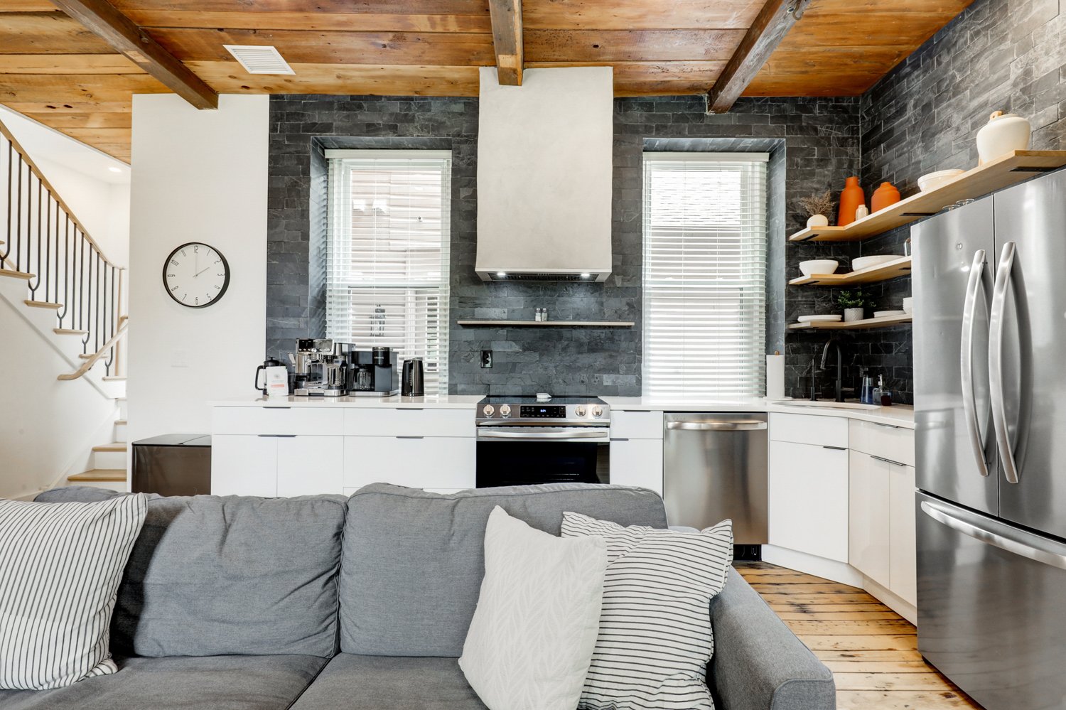 Modern kitchen with dark stone backsplash, white cabinets, stainless steel appliances, open wooden shelves with dishes, and a gray living room sofa with pillows in the foreground.