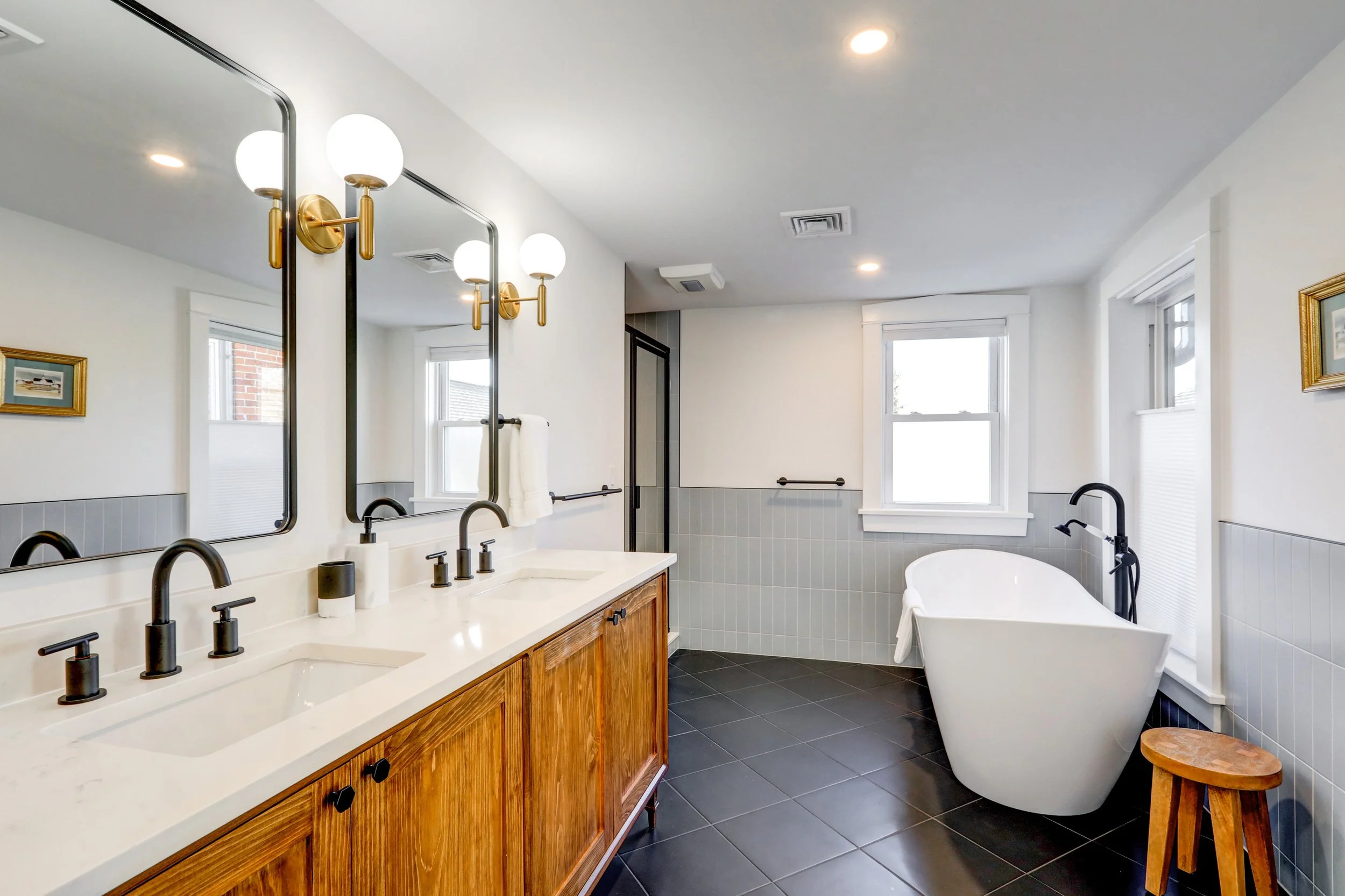 A modern bathroom with a double vanity with black fixtures and a wooden cabinet, a large mirror, and four wall-mounted light fixtures with globe bulbs. There is a bathtub with a black faucet, two windows, and gray tile on the lower part of the walls.