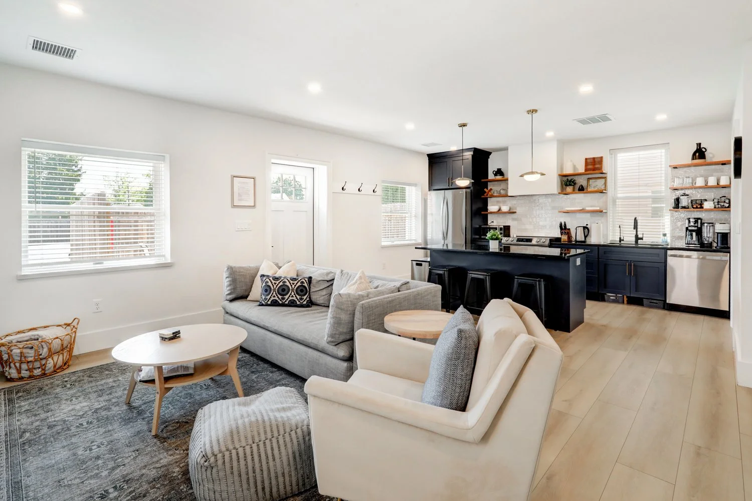 Open-concept living room and kitchen with modern decor, large windows, light wood flooring, white walls, gray sofa, white armchair, coffee table, and navy blue kitchen cabinets with open wooden shelves.