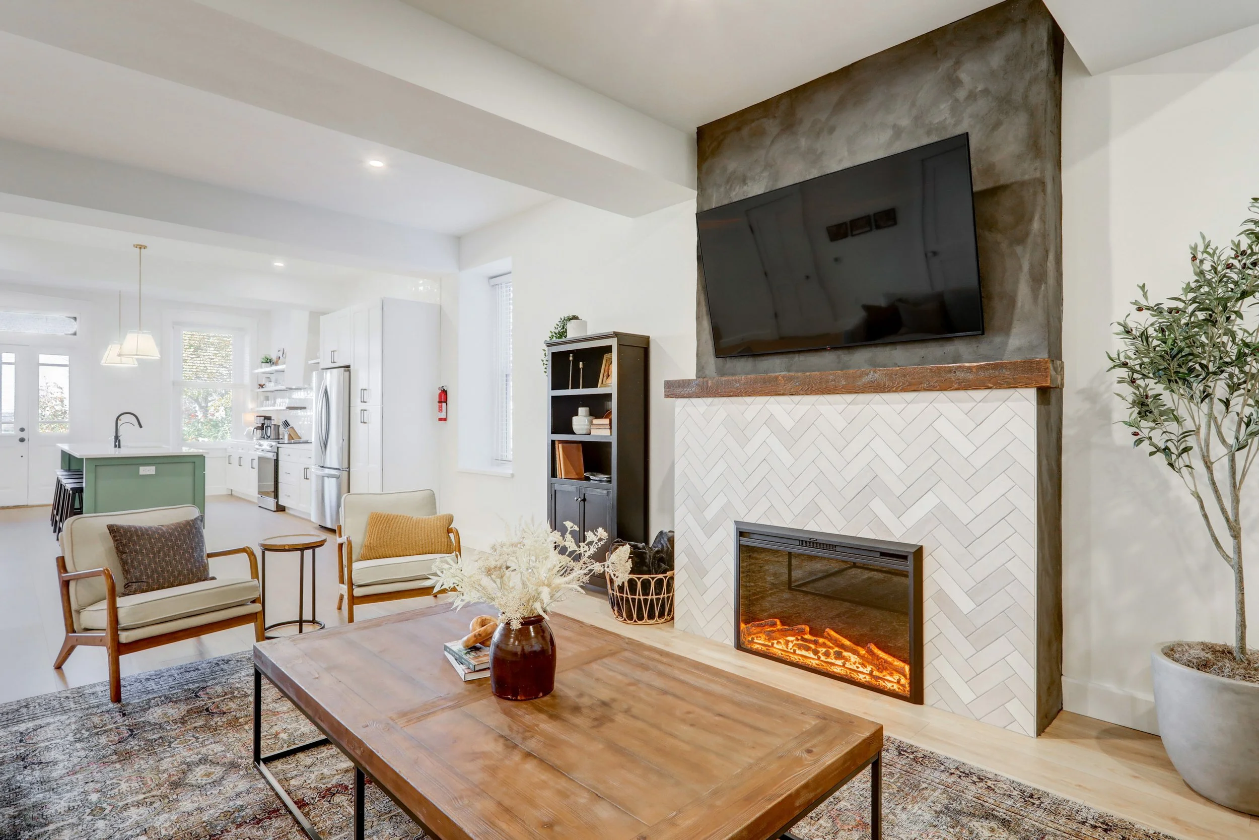 Living room with a modern fireplace, flat-screen TV, wooden coffee table, armchairs, and a large potted plant. Open kitchen area in the background.
