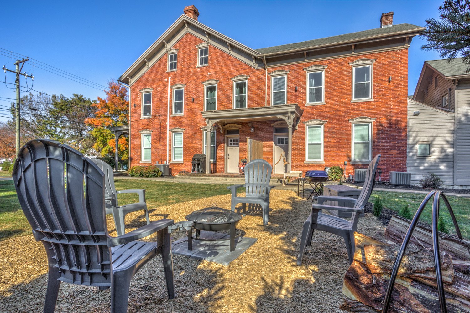 Backyard with lawn chairs around a fire pit in front of a red brick house with multiple windows and a small porch in a suburban neighborhood.