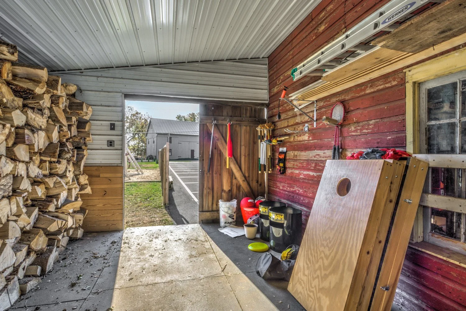 Inside a barn or shed with stacked firewood on the left, tools hanging on the red wooden wall, and various equipment like buckets, helmets, and a large wooden board stored inside. The door is open, revealing a paved area and a metal building outside.