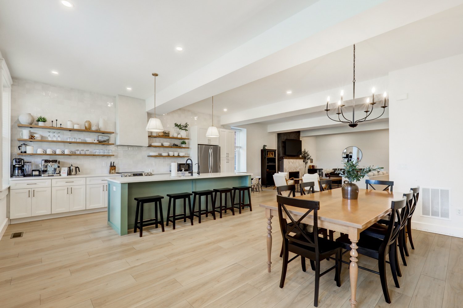 Open-concept kitchen and dining area with a light wood floor, white cabinets, open wooden shelves, a mint green kitchen island with black stools, a wooden dining table with black chairs, and a living area with a fireplace and a chandelier.