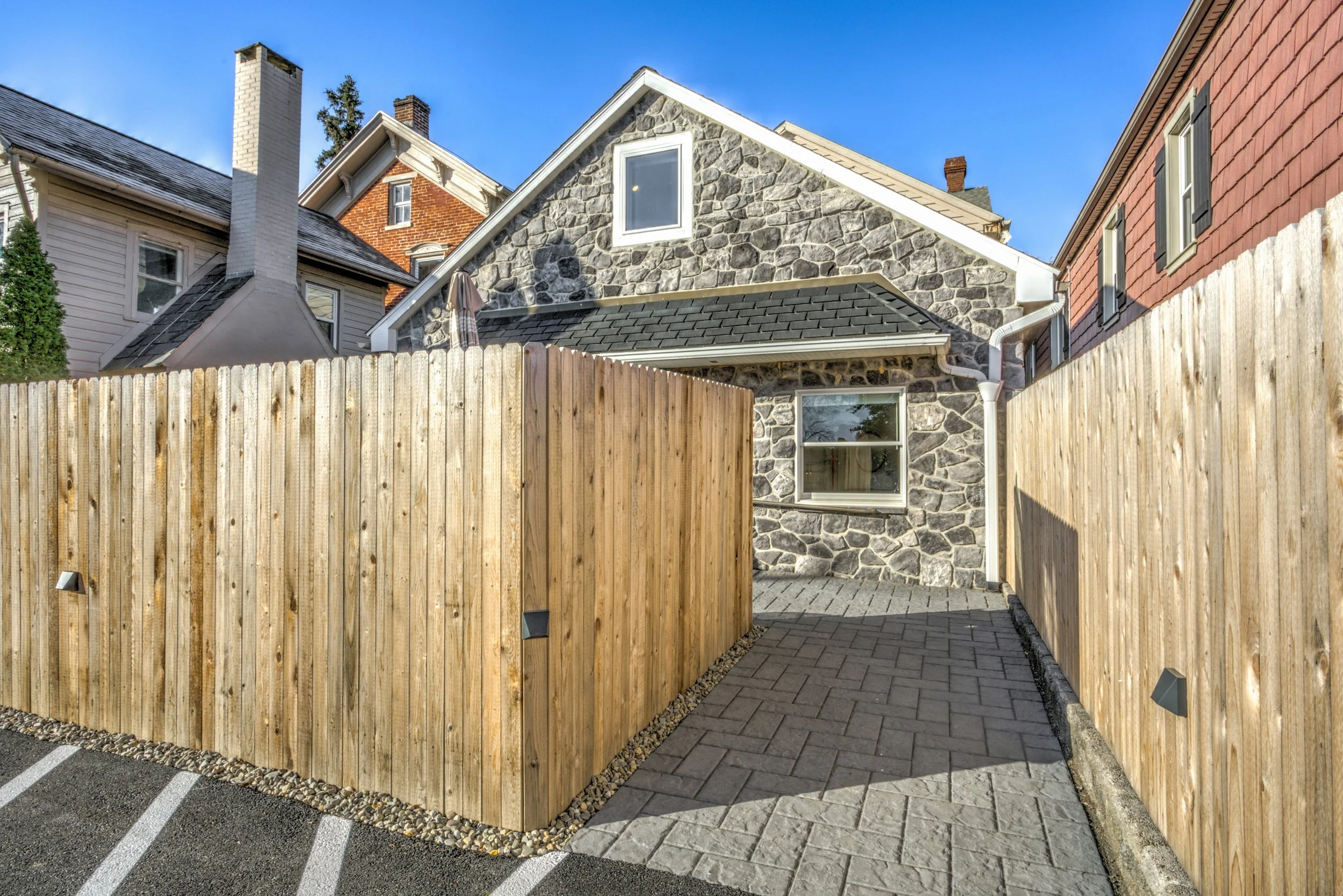 A backyard with a tall wooden fence, a paved pathway, and a stone house featuring gray stone siding and a pitched roof under a clear blue sky.