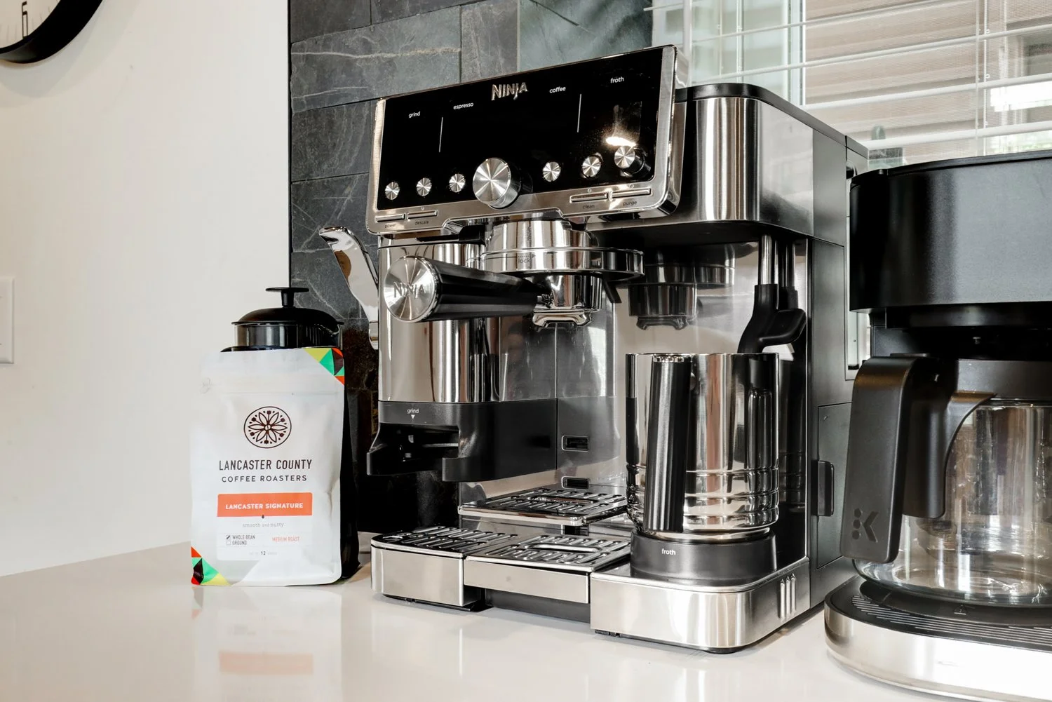 A stainless steel espresso machine on a white countertop, with a bag of Lancaster County coffee roasters coffee and a black coffee grinder to the right.