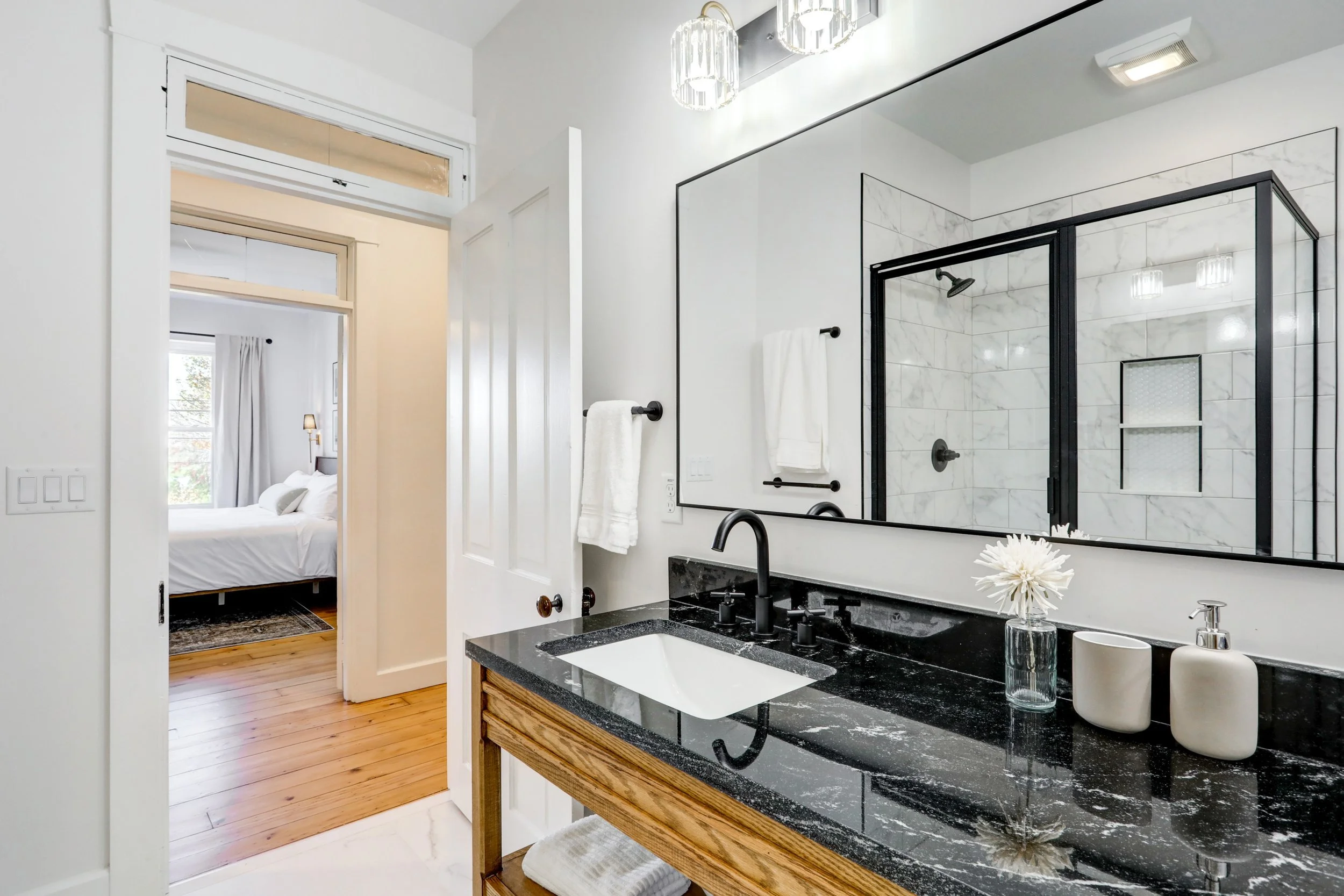 Modern bathroom with black granite sink, white tiles, wall-mounted towel racks, large mirror, and light fixtures. Visible bedroom with bed and window in the background.