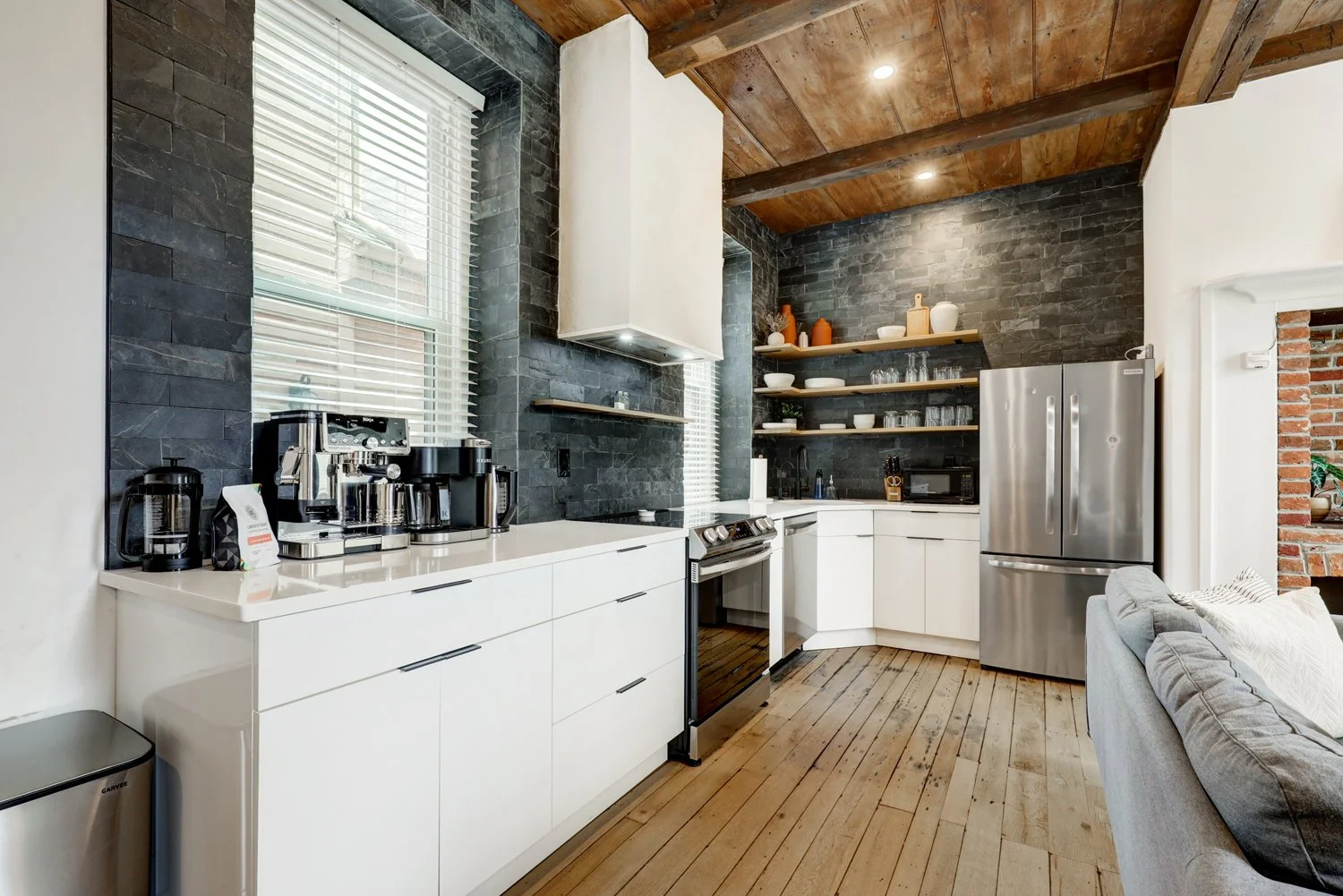 Modern kitchen with black stone walls, white cabinets, stainless steel appliances, open wooden shelves with dishes, and a window with blinds.
