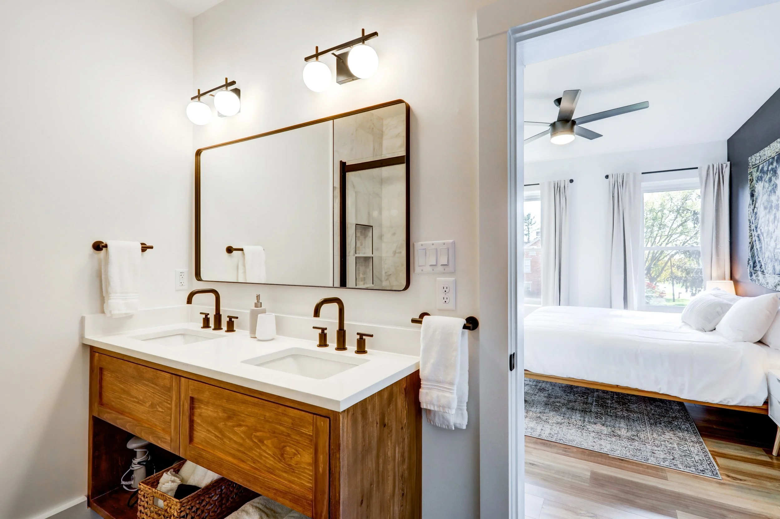 A bathroom with a wooden vanity, a large mirror, two wall-mounted light fixtures, and towels, with a view into a bedroom that has a bed, a ceiling fan, and windows with curtains.