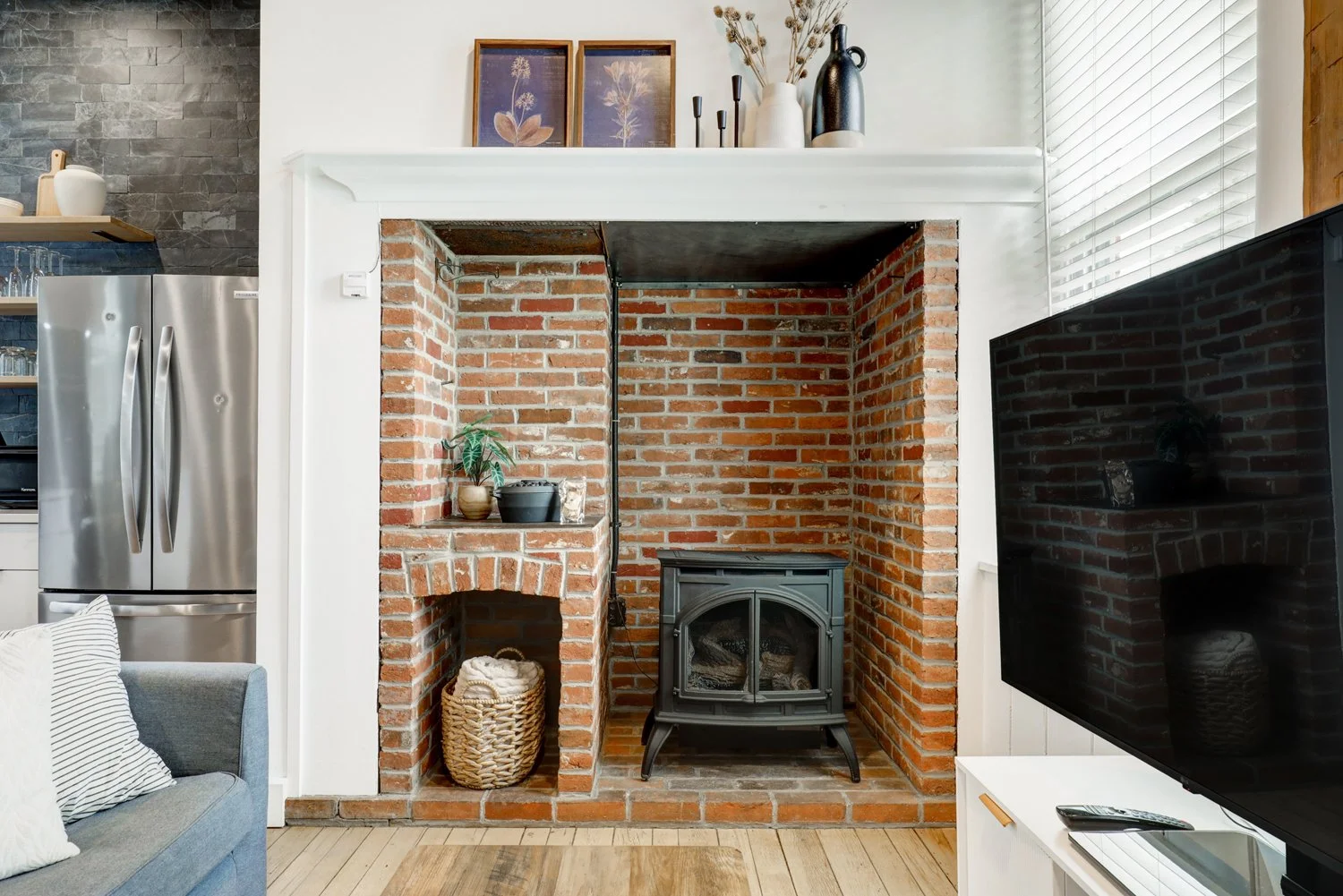 Living room with exposed brick fireplace, black wood stove, decorative plant, baskets, and a white mantle with vases and framed botanical artwork. Part of a gray sofa and a large flat-screen TV on a white stand are visible.