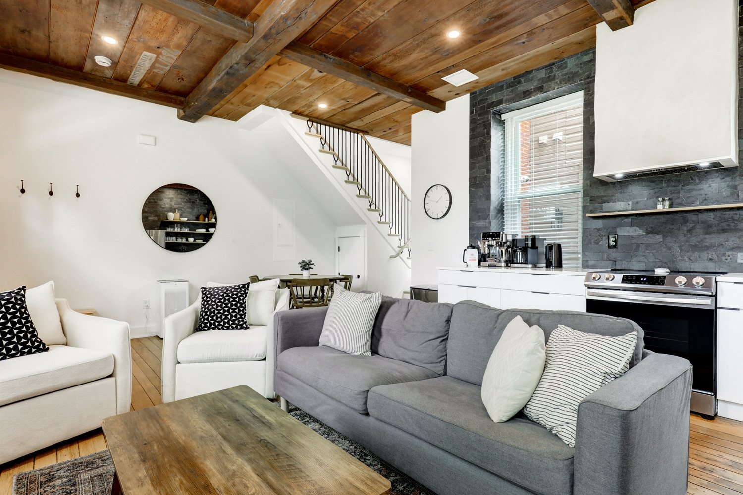 Living room with gray and white sofas, wooden coffee table, black and white pillows, kitchen area with white cabinets, black appliances, stone wall, and a staircase with black railing leading to an upper level.
