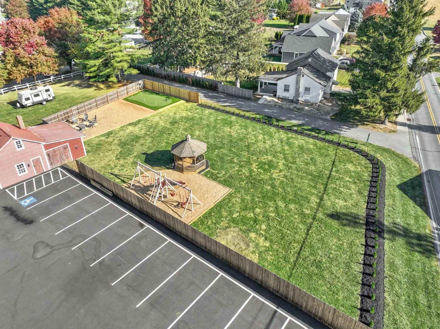A backyard with a grassy lawn, a sandbox with a small gazebo, a swing set, a paved driveway, and a parking lot with one handicap space. There are trees and houses surrounding the yard.