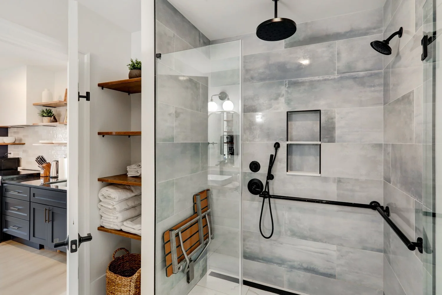 Modern walk-in shower with black fixtures, including a rain showerhead and handheld shower, tiled gray walls, a small wall niche, and a safety grab bar, adjacent to a closet with wooden shelves and towels in a bathroom.
