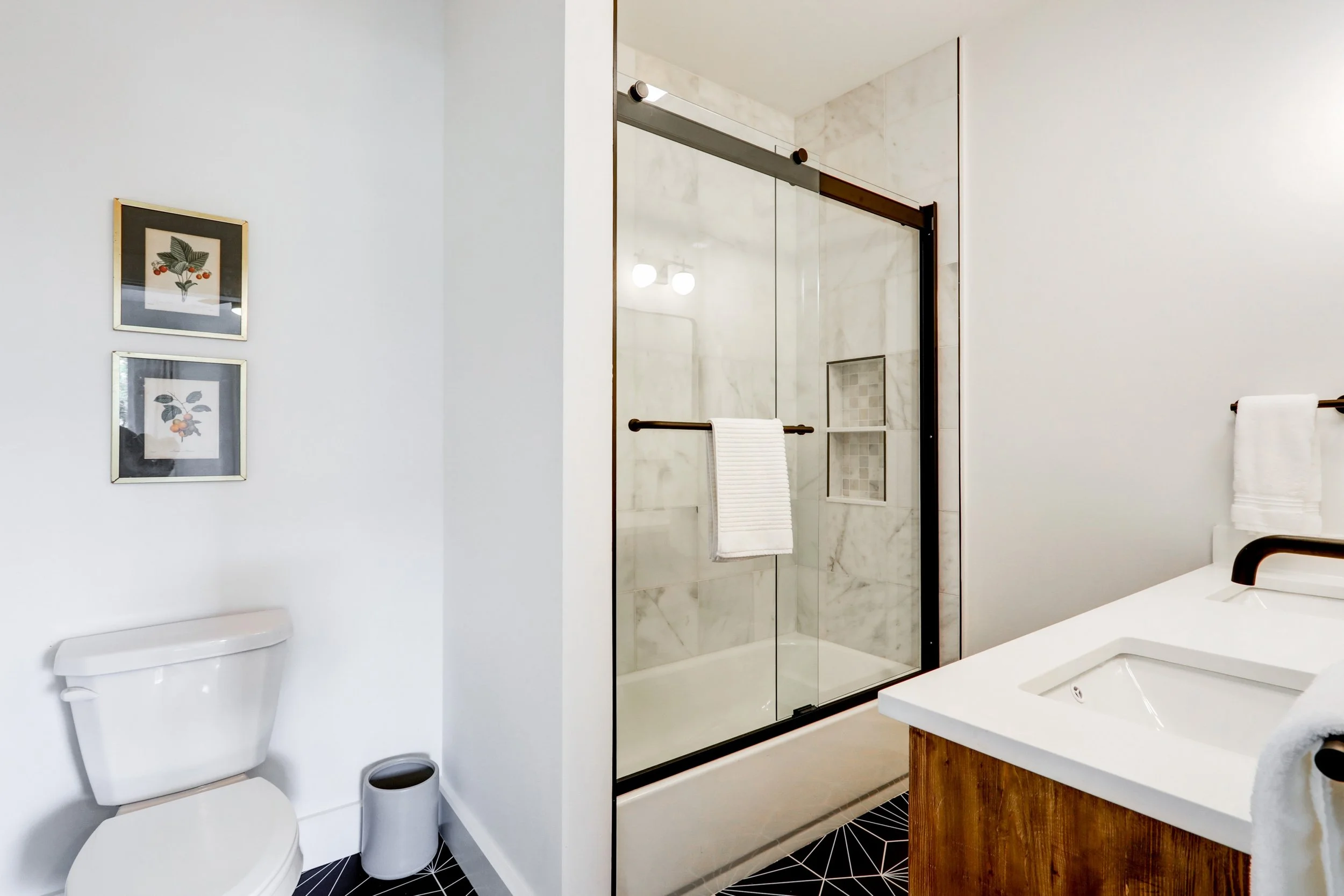 A clean, modern bathroom with a white toilet, framed botanical art on the wall, and a walk-in shower with glass doors and marble tiles.