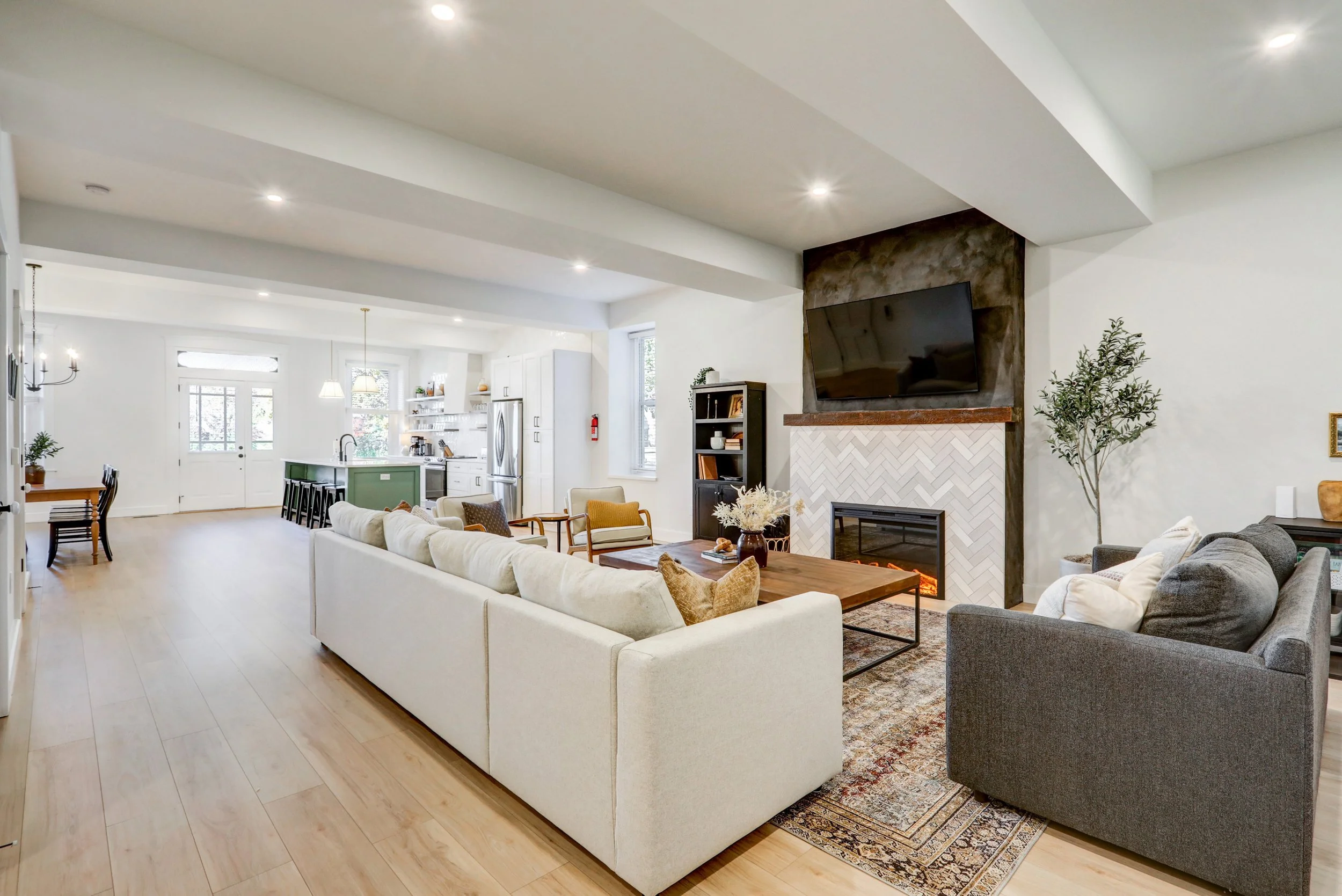 Living room with white sofa, gray armchair, fireplace with a mounted TV, wooden coffee table, and decorative plants, open to a kitchen with an island and bar stools.