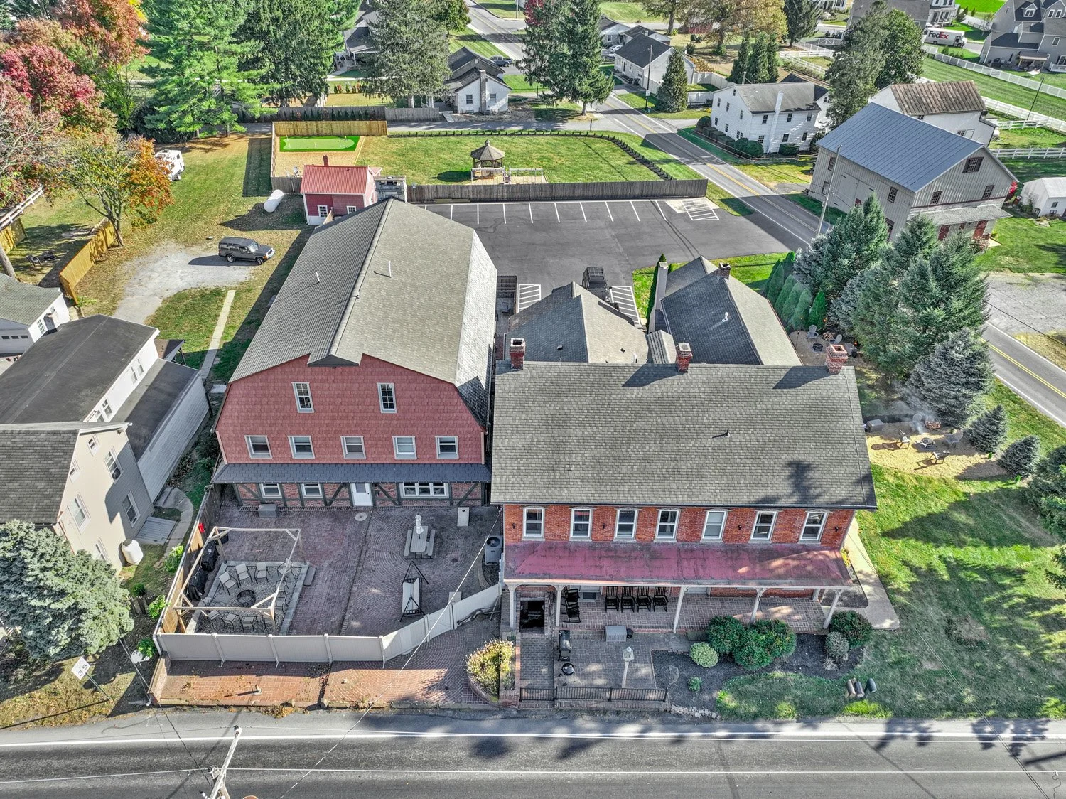 An aerial view of a residential neighborhood showing houses, a parking lot, trees, a fenced backyard with outdoor furniture, a small shed, and a street with power lines.