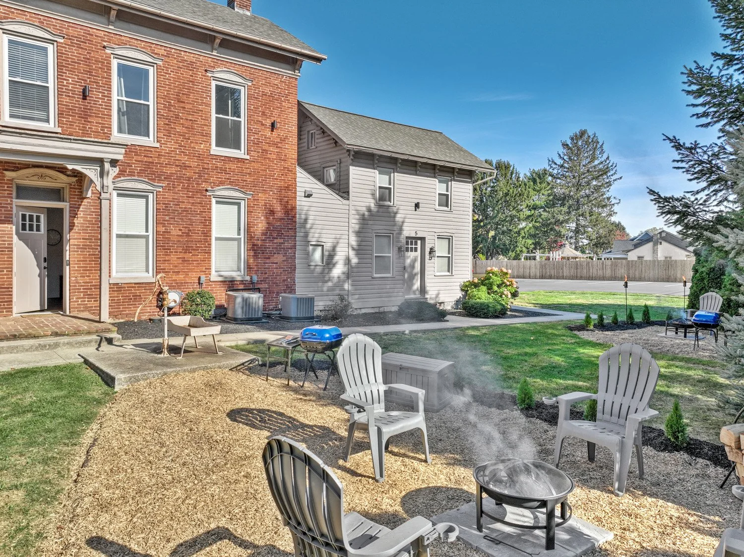 Backyard patio with white Adirondack chairs surrounding a fire pit with smoke, small shrubbery, and a grassy area with a fence. Two houses with brick and light gray siding are in the background under a clear blue sky.